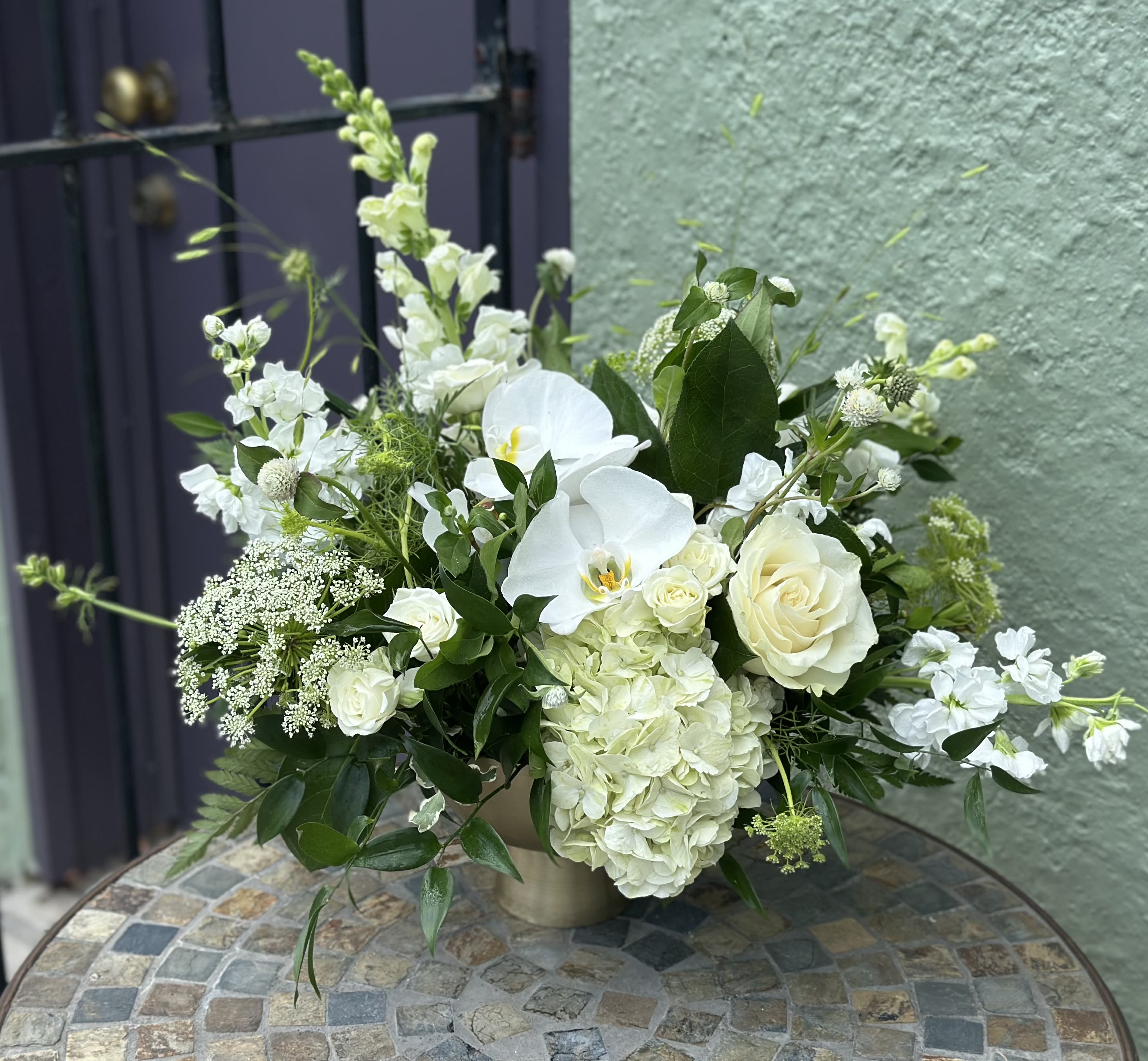 Low white arrangement with roses, orchids, hydrangeas, and snapdragons in a gold vase