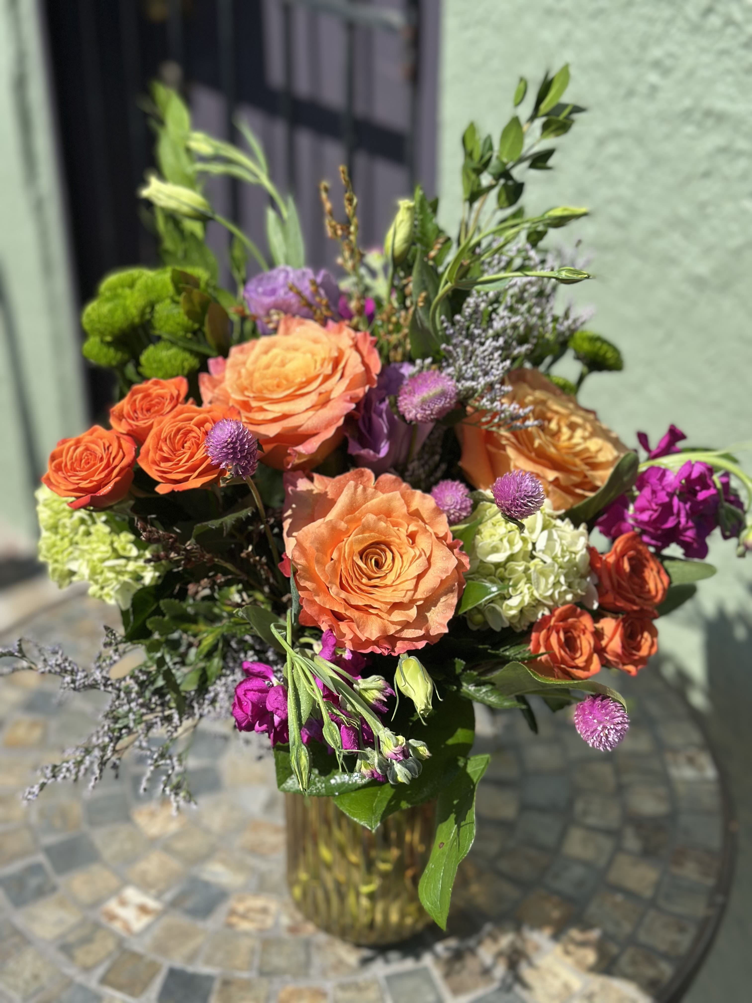 Vase arrangement of bright orange roses, green hydrangeas, and purple accent flowers.