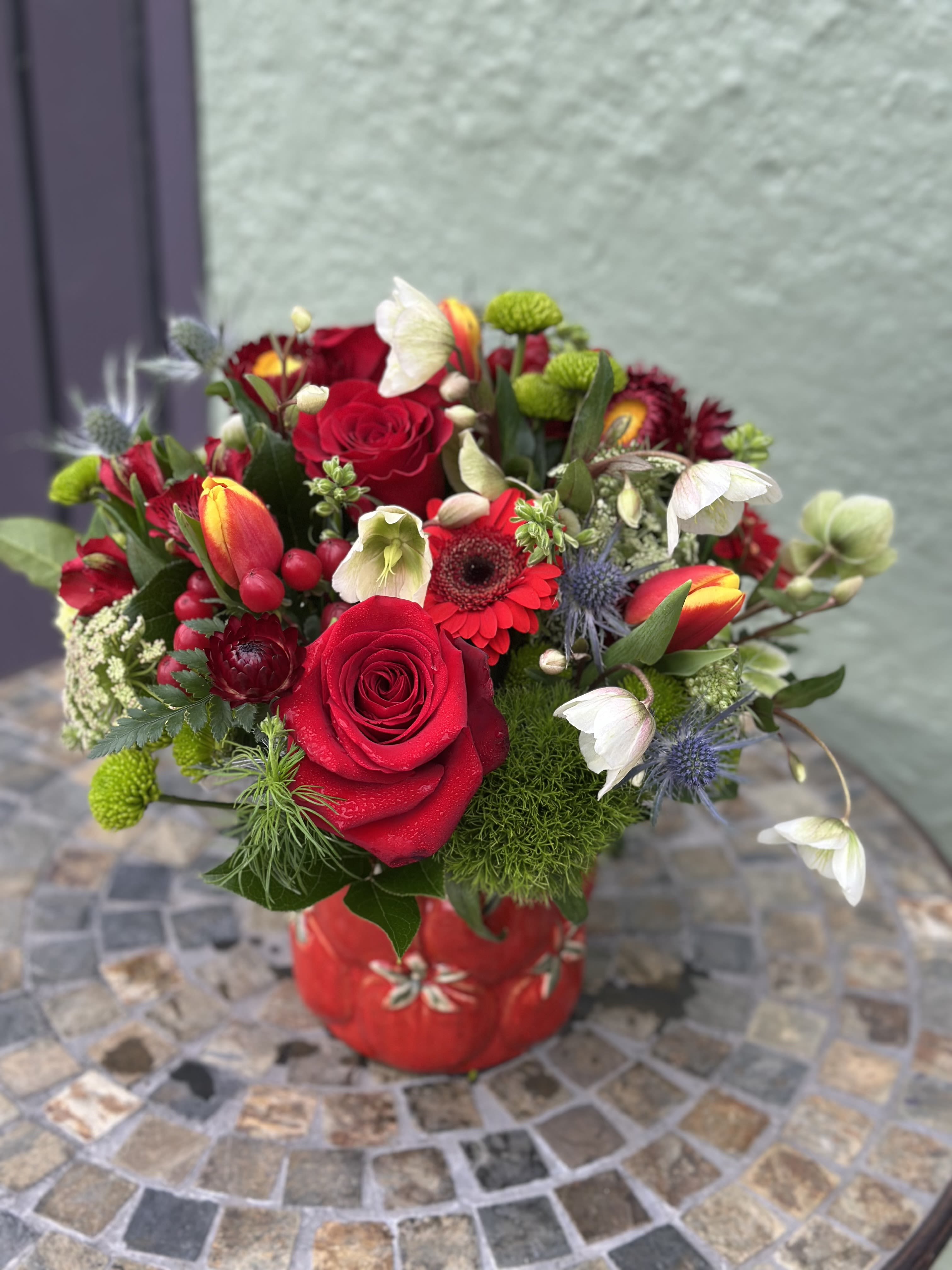 Compact arrangement of red roses, tulips, and mixed blooms in a red ceramic vase on a mosaic table.
