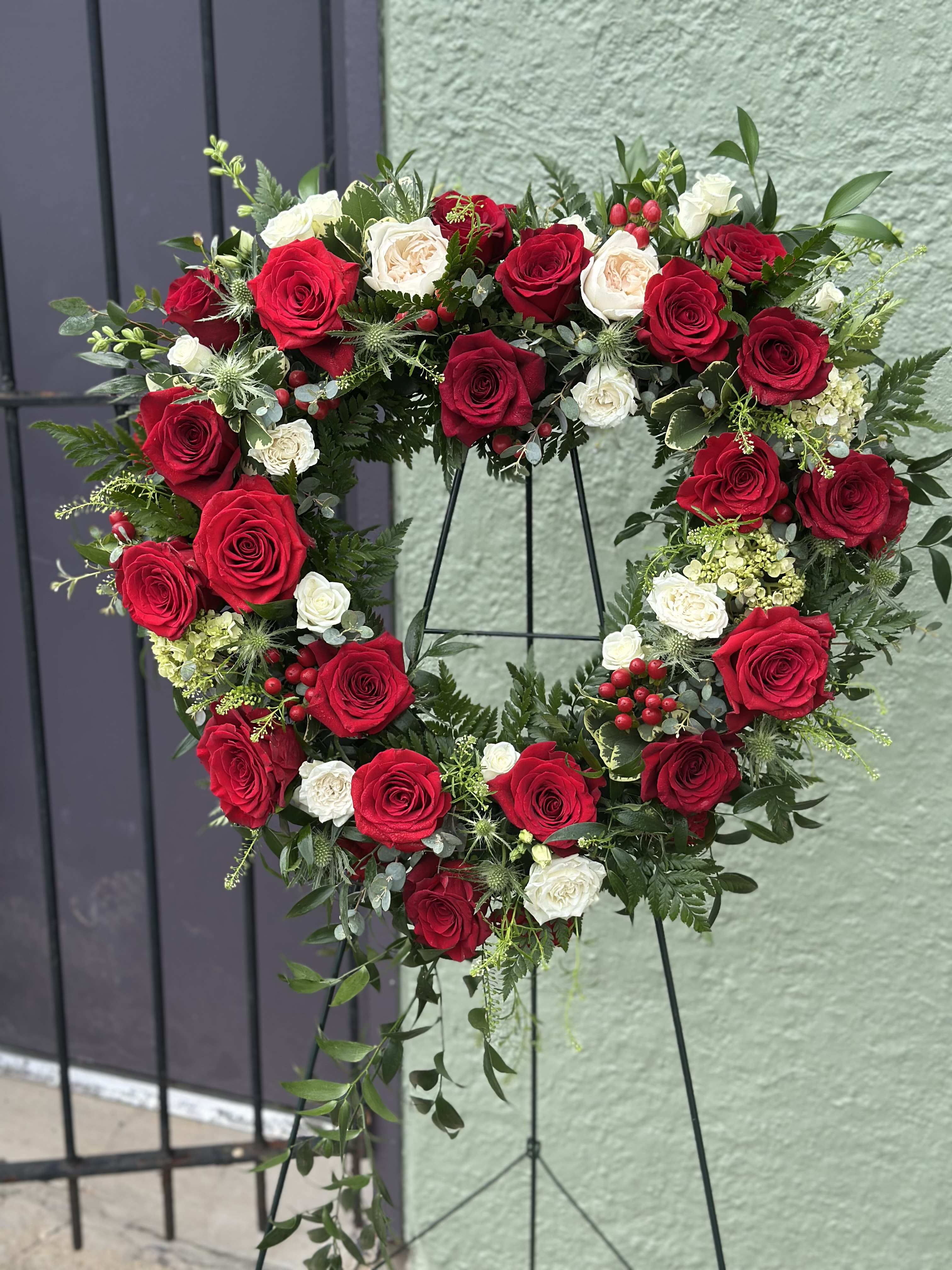Forever Yours - A large(about 24" cross) open heart-shaped floral wreath that speaks of enduring love and reverence - ideal as a sympathy and funeral tribute or a wedding focal piece. This funeral heart arrangement display combines romantic red roses with white and cream roses for contrast, creating a classic red-and-white palette that reads both ceremonial and heartfelt. Mounted on a sturdy black metal easel, the open-center heart makes a powerful visual statement that honors memory or marks a vow.  Carefully layered for full, handcrafted texture, the design also includes pale green hydrangea accents, spray roses, carnations and mixed foliage with small berry-like touches to add depth and subtle color variation. The arrangement is dense and polished, with a balanced mix of blooms and greenery that looks beautiful from every angle and photographs well at a service or celebration. Sized to be prominent but elegant on its easel, it's the kind of tribute that will make the person you're honoring - or celebrating - feel truly seen.