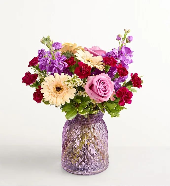 Alluring beauty - From its variety of blooms to its eye-catching crescent shape, this is an out-of-the-ordinary bouquet for that out-of-the-ordinary someone. Roses, Gerbera daisies, and more, in buoyant peaches and purples, arrive lovingly arranged in a lavender vase with cottage charm.
