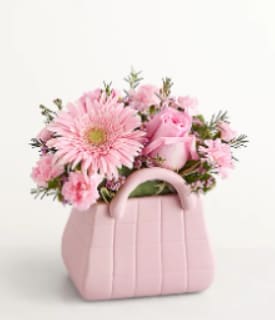 Designer's Dream Bouquet - A cheerful cluster of soft pink gerbera daisies, roses, and carnations arranged in a pastel pink handbag-shaped vase makes a playful bouquet for any occasion that feels like a present already. The large gerbera face draws the eye while the rounded silhouette and gentle palette create a warm, romantic tone-perfect for a mom, sister, or close friend who should feel celebrated and seen.  This compact, full arrangement uses light greenery and delicate filler blooms tucked between the larger blossoms for texture and a polished finish. The pink handbag-style container doubles as a keepsake decorative piece, so the recipient gets flowers plus a charming accent for a vanity or tabletop. Sized to sit comfortably on a desk or small table, it's an easy, everyday way to say "thinking of you" or "happy birthday."