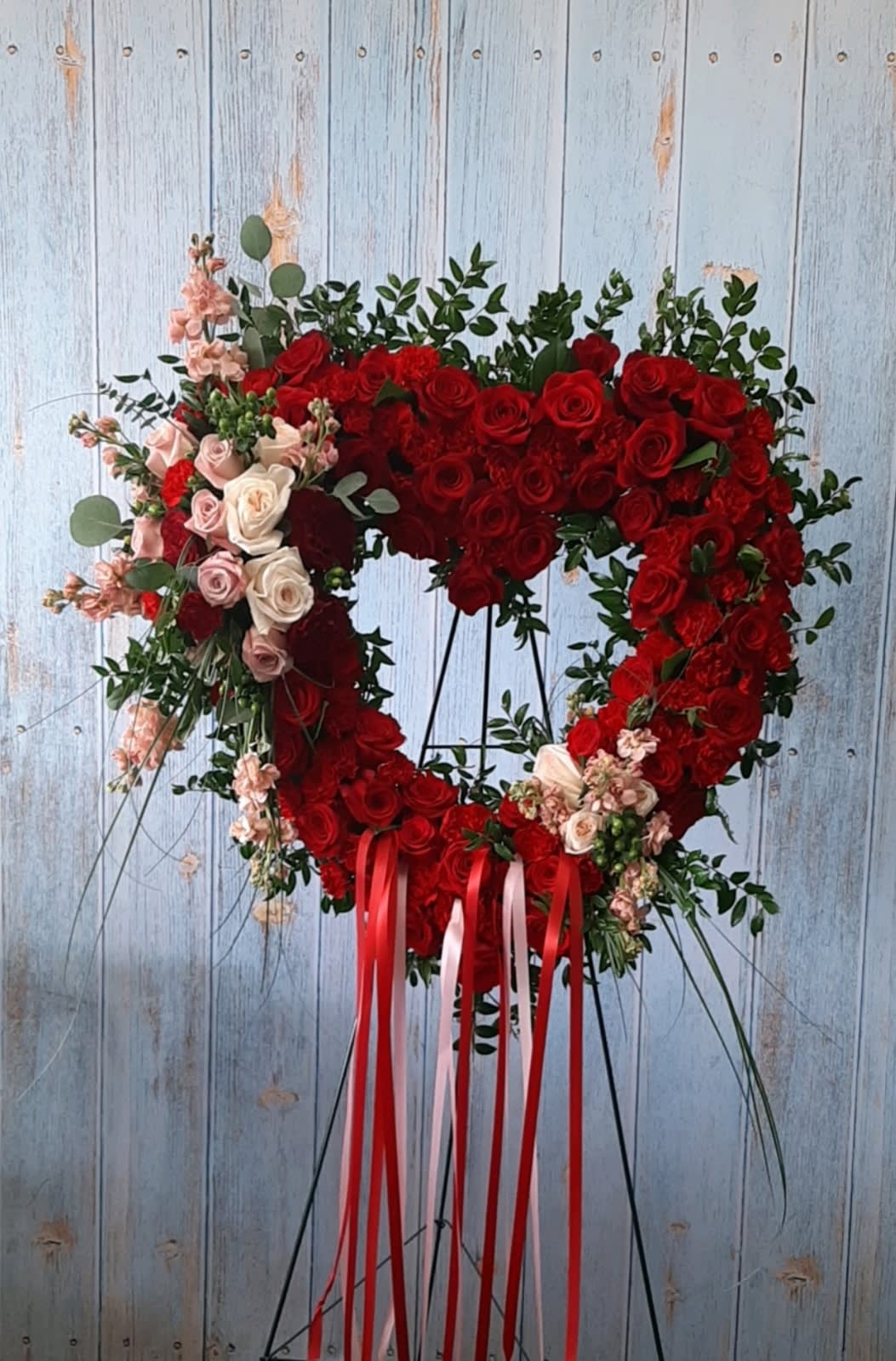 Heart-shaped red rose wreath on a stand with pink accents and ribbon tails