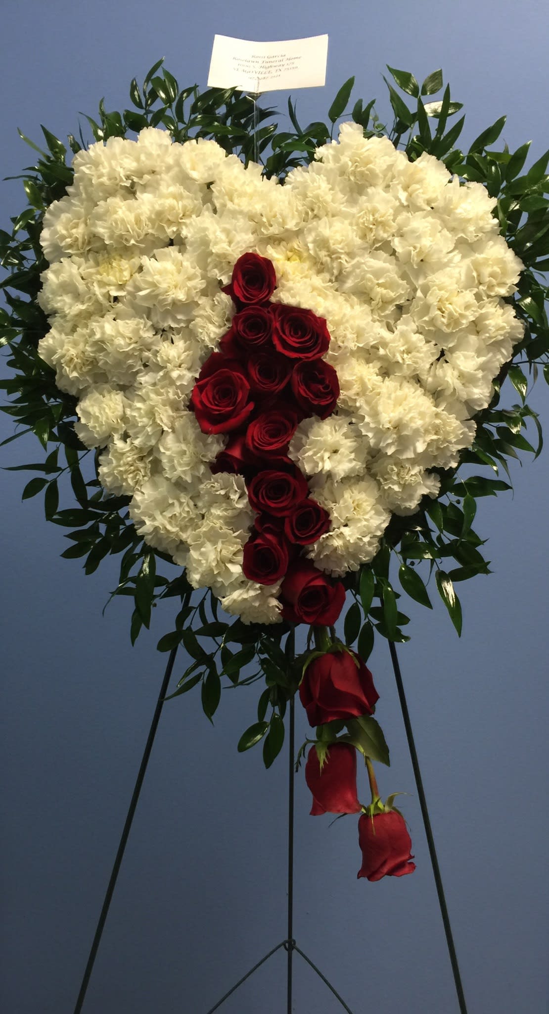 Heart-shaped standing spray of white carnations with cascading red roses on an easel.