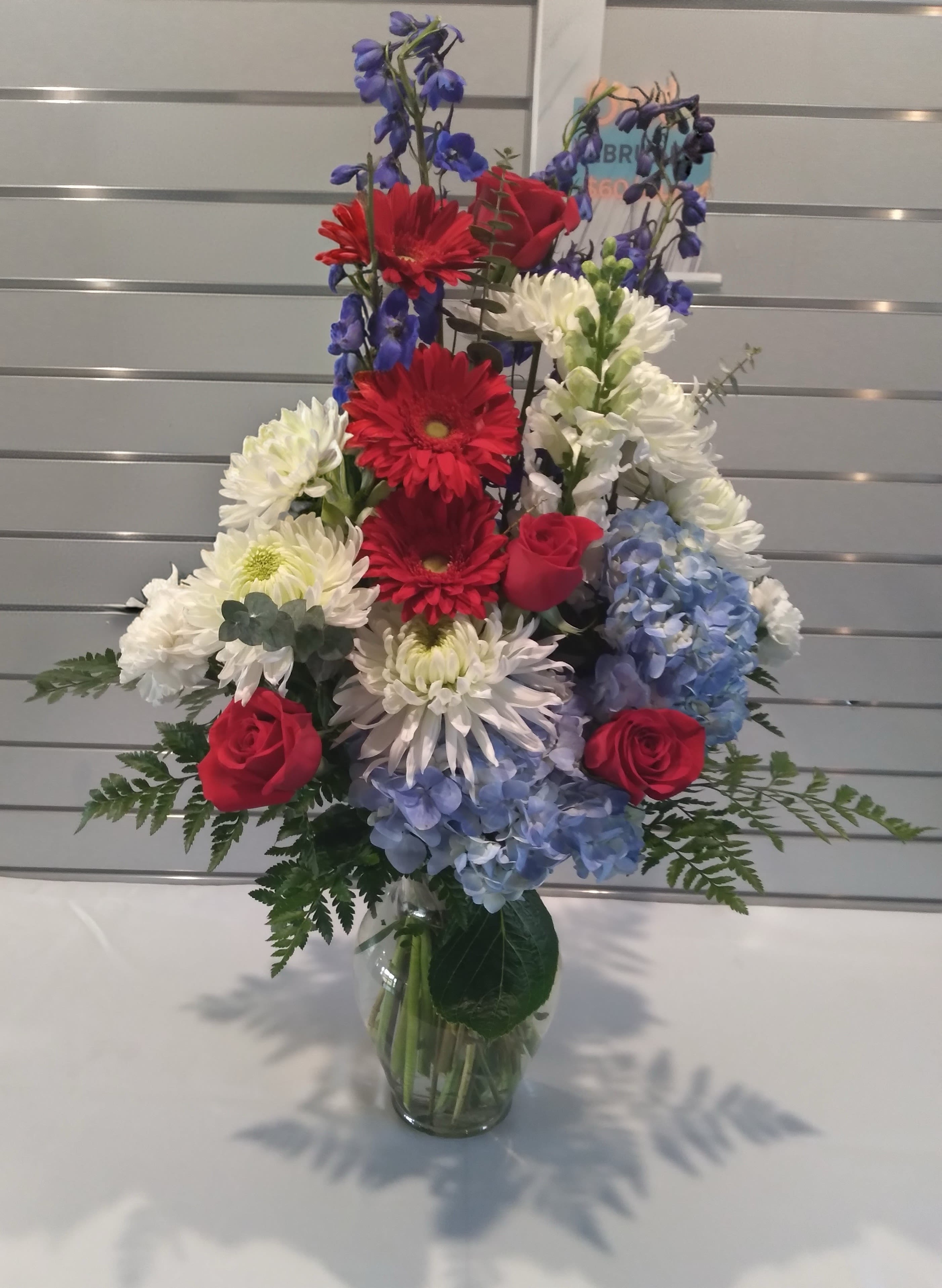 Sam - A tall, layered mixed flower arrangement made for celebration and everyday display, designed to make the person you're thinking of feel noticed and honored. This mixed-color flower arrangement is an excellent choice for a patriotic holiday, anniversary, or a bold thank-you bouquet-its red roses, white daisy-like blooms, and blue hydrangea create a striking, classic contrast that reads festive without feeling gimmicky.  The arrangement sits in a clear glass vase and features upright blue delphinium spikes that add vertical rhythm and drama above fuller blooms, with fern-like foliage fanning at the base for texture and balance. Expect vivid red roses, clusters of blue hydrangea, white daisies, and complementary seasonal stems arranged to show off each flower's color and form-the result is visually rich from every angle.  Perfect for gifting to a friend, family member, or colleague for holidays, celebrations, or just because, this design brings a polished, uplifting presence to any room. The clear glass vase keeps care simple-fresh water and a trim every few days will help the blooms stay lively-and available in a tall, showy presentation that works well on mantels, dining tables, or as a centerpiece.