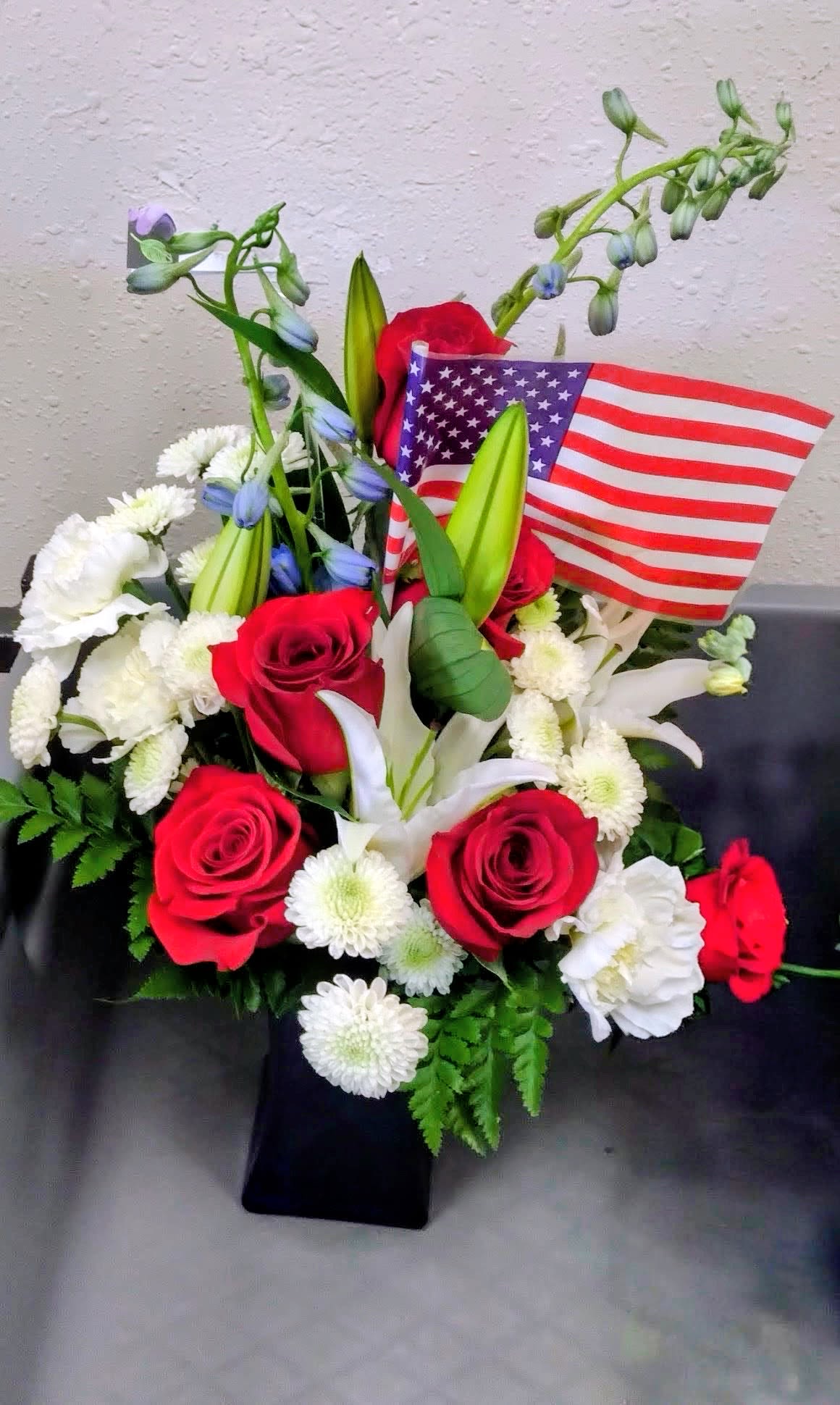 Liberty - Bold red roses with crisp white daisies and a few elegant white lilies create a striking patriotic flower arrangement perfect for Memorial Day, Independence Day, Veterans Day, or any occasion calling for a respectful tribute. Tall blue-tinted accent stems lift the composition and add celebratory height, while the American flag tucked at the back makes the red, white, and blue message unmistakable - the kind of arrangement that will make the person you're honoring feel remembered and respected.  Gathered in a sleek black vase, this formal design mixes classic roses and lilies with daisy-like blooms and lush greenery for contrast and texture. The taller accents give visual movement and make the piece suitable for display on a memorial table, as a center on a reception counter, or beside a gravesite. Simple care: keep water fresh and placed out of direct heat to extend vase life and preserve the arrangement's crisp colors.