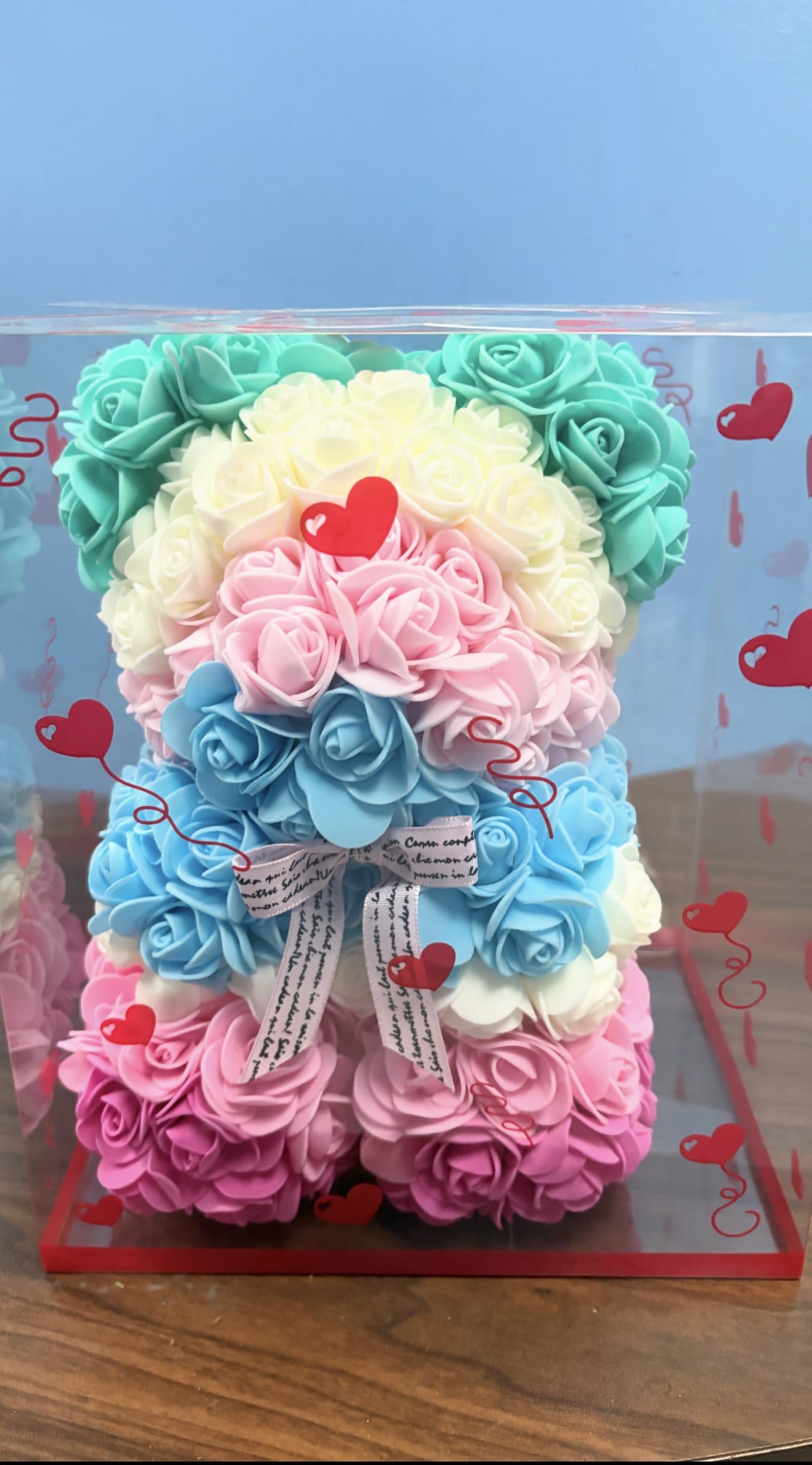 Pastel Petals Bear - Soft pastel rose blooms sculpted into a playful teddy-bear shape make this boxed floral keepsake an instant smile-maker for birthdays, anniversaries, baby showers, or Valentine's Day. The rose-shaped silk flowers in pale pink, sky blue, white and aqua create a whimsical, love-themed presentation that reads as both romantic and gently celebratory - the perfect way to make the person you're thinking of feel seen and adored.  Tightly packed silk roses form a compact, rounded figure (teddy-bear/heart-inspired) and arrive enclosed in a clear plastic display box printed with red heart graphics. A printed ribbon bow and heart decorations finish the gift-ready look; clear plastic wrap and decorative stickers keep the design neat and display-ready on arrival. Lightweight and long-lasting, this keepsake-style arrangement requires no water and is meant to be enjoyed as a decorative memento for years.