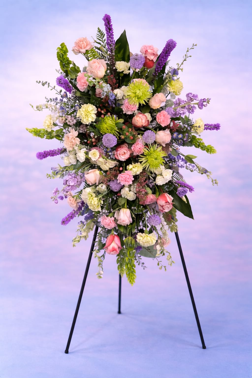 Whispers of the Garden - Whispers of the Garden is a graceful sympathy spray designed to reflect a life remembered with tenderness and love. Featuring soft blush roses, delicate lavender blooms, fresh green chrysanthemums, and airy seasonal accents, this arrangement brings together soothing pastels and natural textures in a beautiful tribute.  The flowing design and varied blooms create a gentle, garden-gathered feel — symbolizing peace, renewal, and the quiet comfort of cherished memories. Thoughtfully arranged on a standing easel, this piece offers a serene presence at funeral services or celebrations of life.  Its soft purples represent admiration and dignity, blush tones express gratitude and love, and fresh greens symbolize renewal and everlasting remembrance.