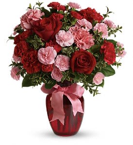 Red and pink roses and carnations arranged in a red glass vase with a pink ribbon bow