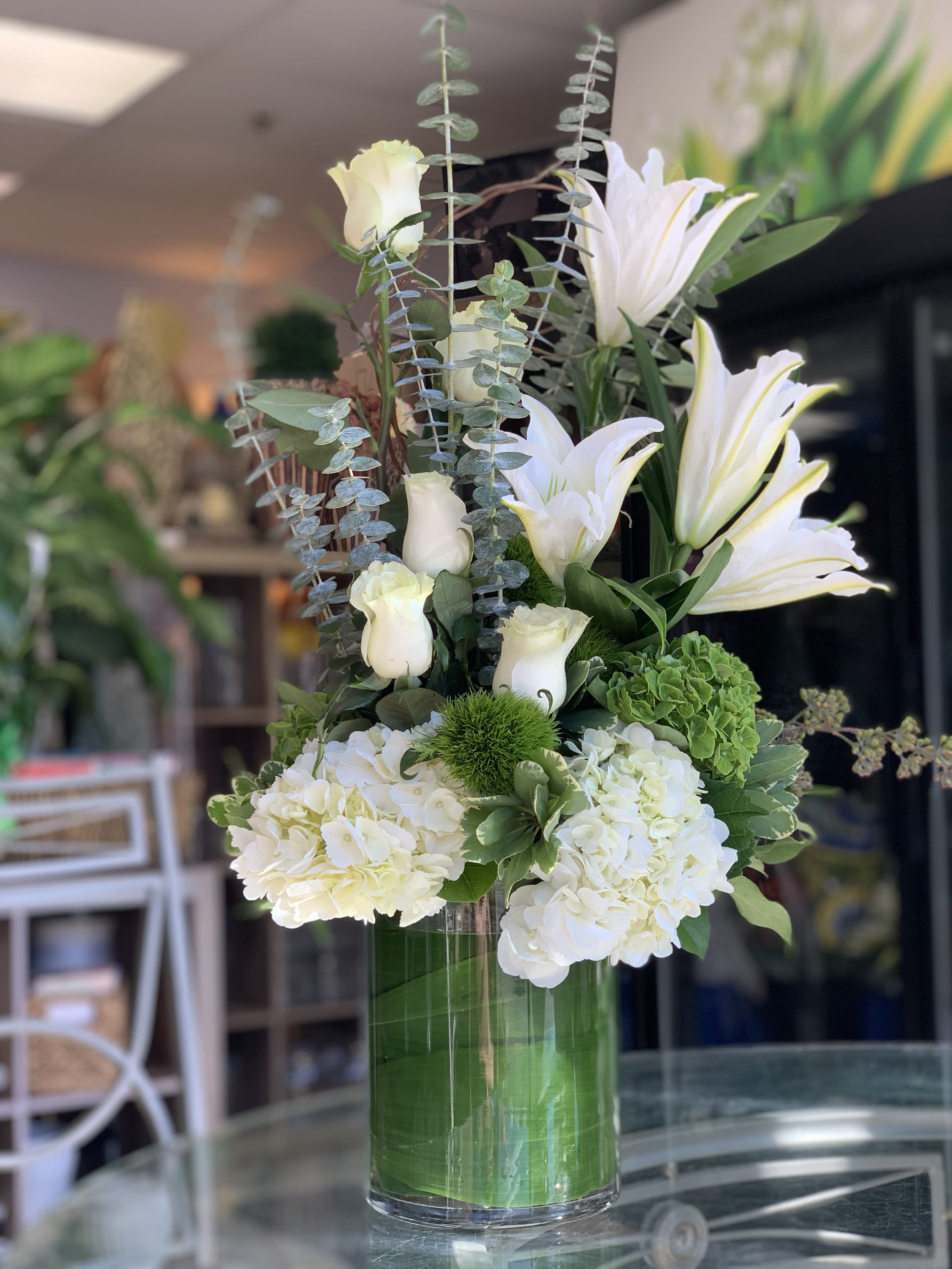 Tall white arrangement with lilies, roses, and hydrangeas in a clear green-lined glass vase.