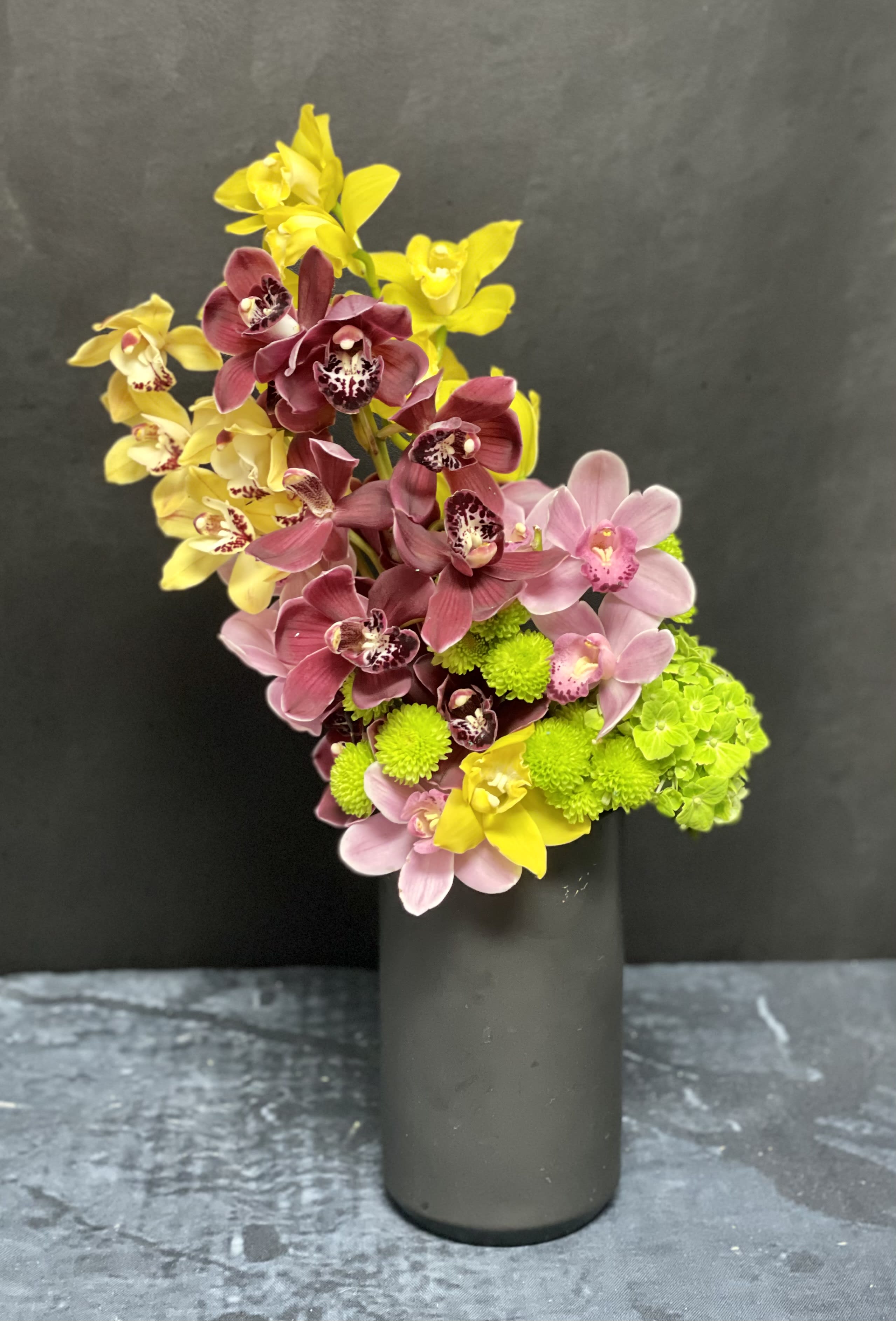 Tall black vase with pink and yellow orchids and bright green blooms in a modern angled arrangement