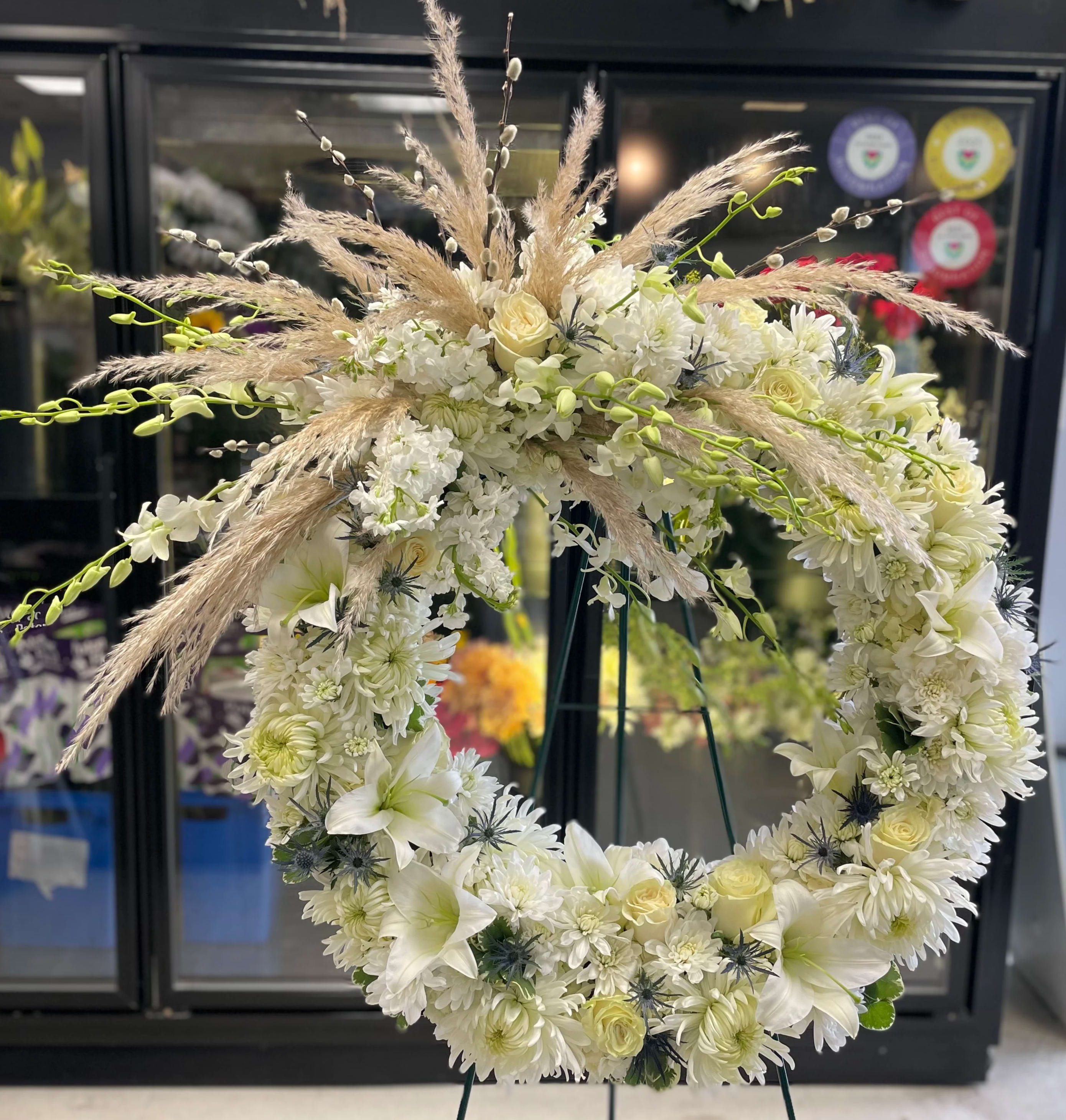 Always Remembered - This large classic wreath is delivered on an easel, and is a thoughtful expression of sympathy and admiration. A standing wreath created from fresh white flowers such as roses, lilies, white orchids, white stock, blue thistles and cushion spray chrysanthemums and accented with greenery. 