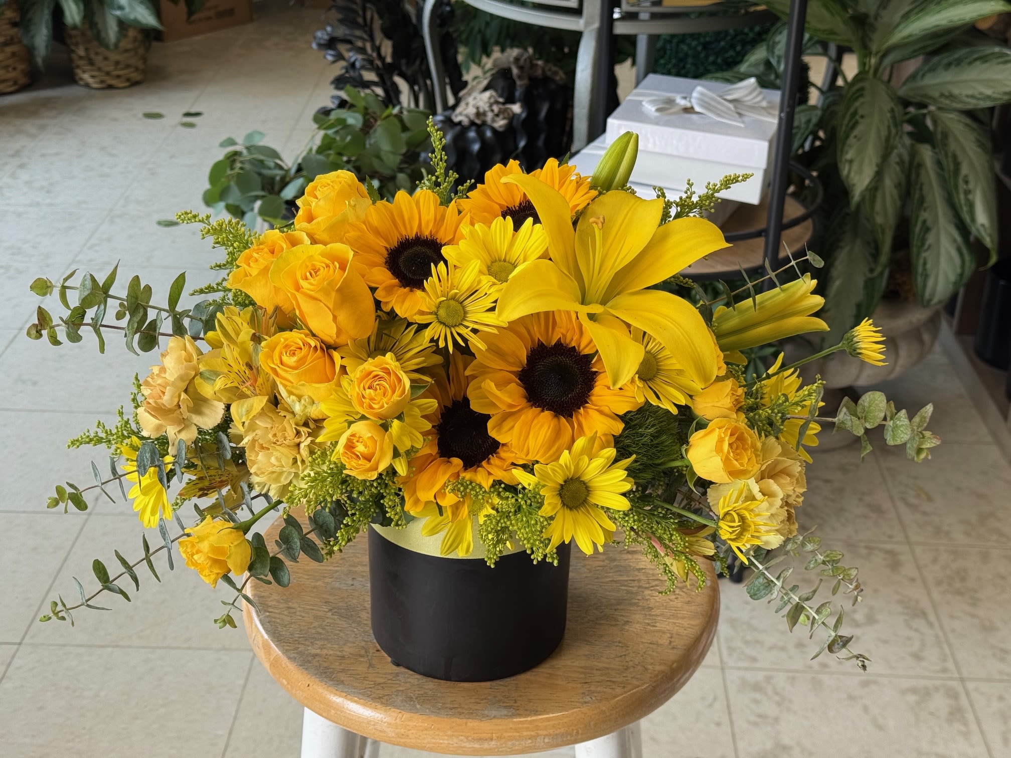 Bright yellow arrangement of sunflowers, lilies, roses, and daisies in a black cylinder vase