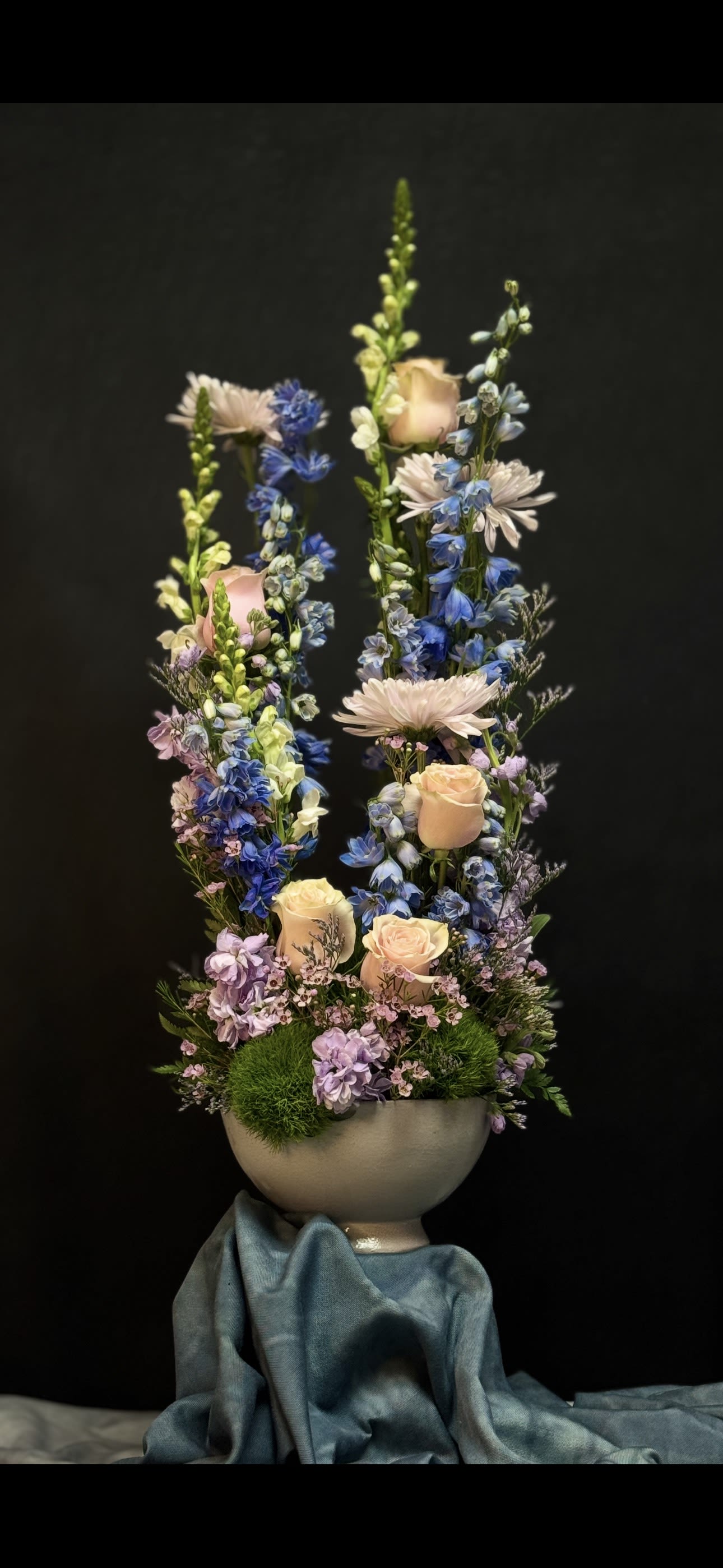 Tall pastel arrangement of peach roses and blue delphinium in a white bowl vase on draped fabric
