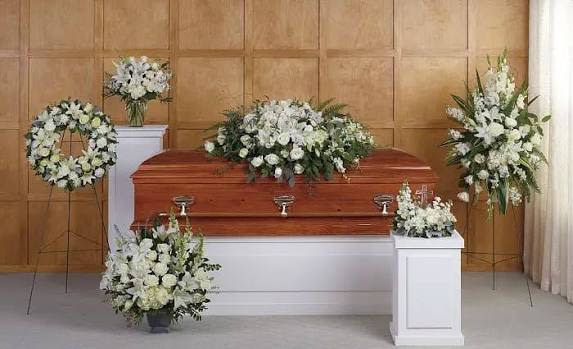 Petal Perfection’s White Solace Collection  - A serene all-white presentation designed to honor and comfort-the White Solace Collection features coordinated funeral and sympathy arrangements surrounding a polished wooden casket to create a calm, respectful setting. This funeral flower arrangement group is ideal for a memorial service or celebration of life, conveying peace and sympathy with an elegant, monochromatic palette that lets the sentiment shine through.  Pure white roses, lilies, carnations, and chrysanthemums form the low, full casket spray with rounded, soft textures that sit gracefully atop the casket. Matching tall standing sprays and a circular wreath on metal easels echo the same formal styling, while additional arrangements in a clear glass vase, low containers, and white display pedestals provide balanced height and visual flow around the space. The mix of blooms offers gentle contrasts of shape and scent while maintaining a unified white-and-green color story.  Arranged to comfort the family and honor the loved one, these pieces read as cohesive and thoughtfully planned-she'll feel remembered and respected by this quiet, dignified tribute. Available as a coordinated group or as individual pieces to suit the service size; please contact us for delivery, setup on-site, or to confirm dimensions and placement for your venue.