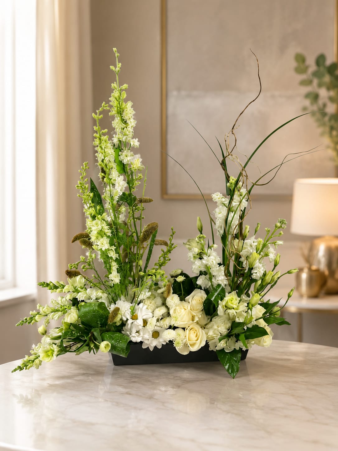 Petal Perfection’s Eternal Grace - A serene, contemporary centerpiece that reads as both refined sympathy arrangement and an elegant gift for Mother's Day, an anniversary, or a thoughtful get well bouquet. This low rectangular black container holds a densely clustered cushion of white roses, spray roses, daisies, hydrangea, and stock, while taller snapdragons, stock stems, and sculptural greenery rise at the sides to create an airy, asymmetrical silhouette. The white-and-green palette feels calm and timeless-perfect for Easter, weddings, memorials, or simply to bring quiet beauty to a dining table or office console.  The mix of soft round blooms and linear green accents gives pleasing contrast in texture: lush hydrangea and roses at the base, delicate daisy centers for brightness, and taller snapdragons and curly grasses that add movement and modern height. Arranged in a sleek black rectangular vessel, the piece measures to suit tabletops and reception desks and reads well in minimalist or formal settings. It's the kind of floral gift that makes the person you're thinking of feel genuinely seen-appropriate for birthdays, sympathy, housewarmings, thank-you gestures, graduations, or "just because" moments. For care, keep water fresh and in bright, indirect light to extend the life of the flowers.