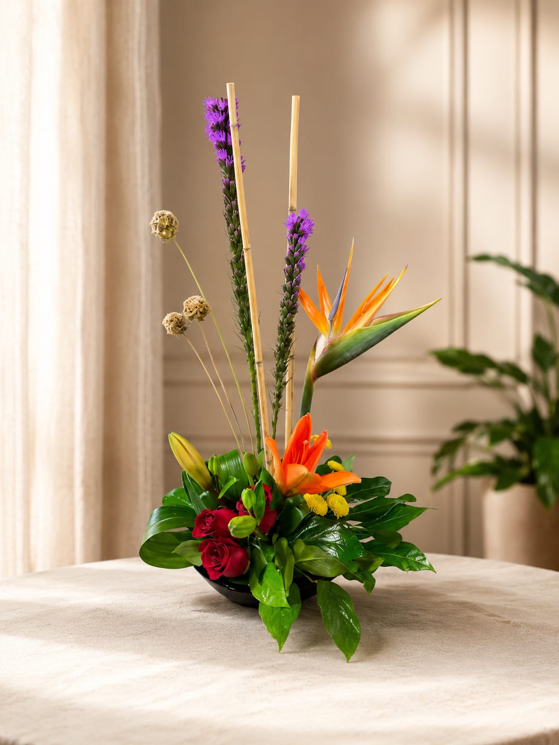 Petal Perfection’s Tropical Delight - A bold vertical statement that brings tropical energy to any celebration, this flower arrangement is perfect as a Mother's Day bouquet, a birthday surprise, or a vibrant housewarming gift. The design pairs tall purple flower spikes and slender decorative sticks with a vivid bird of paradise and an orange lily for a striking focal line, while deep red roses tucked low add warm romantic depth-ideal for anniversaries, Valentine's Day, or to say "Thinking of You" with flair.  Set in a sleek black ceramic bowl, glossy dark green foliage forms a dense, rounded base that anchors the composition and lets the bright reds, oranges, yellows, purples, and touches of blue pop against the container. The arrangement's compact footprint makes it easy to place on tabletops or entry consoles; the tall vertical elements give it gallery-like presence without overwhelming a space.  Send this as a cheerful get-well bouquet, a festive addition for Easter or summer gatherings, or "just because" to make someone feel truly seen-the person receiving it will notice the artistry and bold color. Care is simple: keep water fresh, trim stems as needed, and display away from direct heat to extend the life of the tropical blooms.