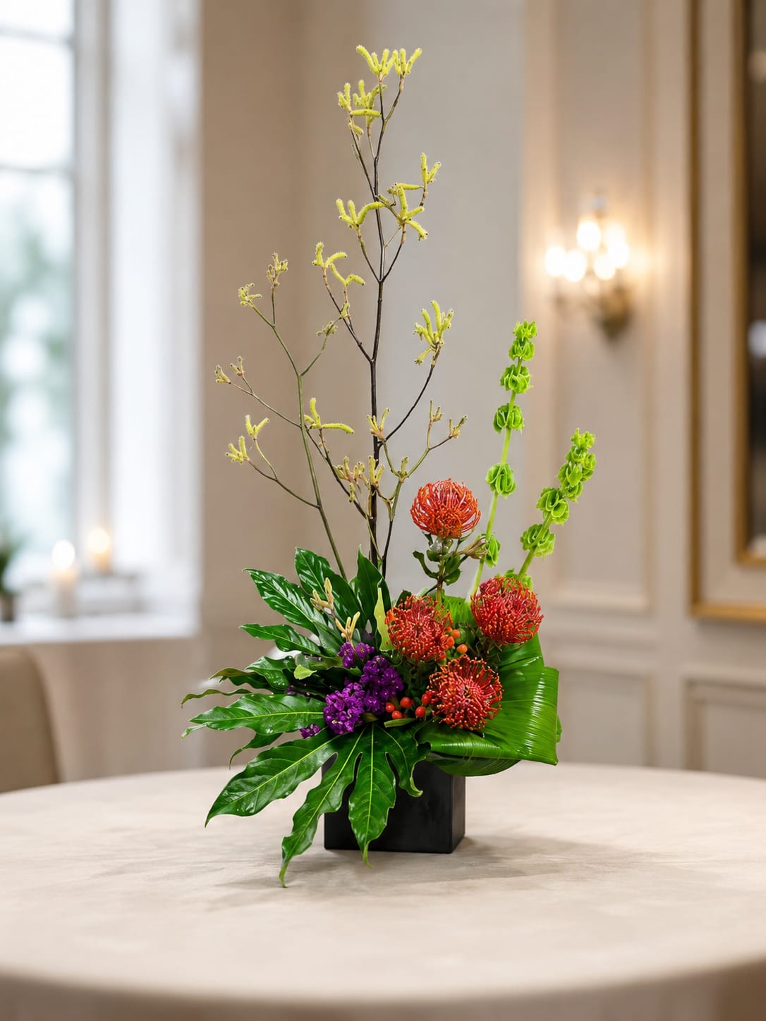 Petal Perfection’s Pretty Protea - A sculptural statement for Mother's Day, a modern housewarming gift, or a bold anniversary surprise: this flower arrangement centers on red pincushion protea that form a textured focal cluster, their dramatic form softened by small purple blooms and red berries. Tall branching stems of small yellow blooms rise above the compact base while vertical green Bells of Ireland create a striking architectural line, giving the piece a contemporary, asymmetrical profile that reads like floral art.  Set in a sleek black rectangular ceramic container, glossy tropical foliage fans outward to balance the composition and create negative space for a minimalist, refined mood. Colors blend red, pink, purple, yellow, orange and green for a vibrant mixed-color palette that suits spring, summer or any celebration-birthdays, graduations, thank-you gestures, get-well wishes, or just-because moments. Dimensions are intentionally compact with tall vertical accents for visual impact without overwhelming tabletops or office desks; care is simple-keep fresh water in the container and trim stems for longer enjoyment-so the person you're thinking of will feel seen, celebrated, and delighted.