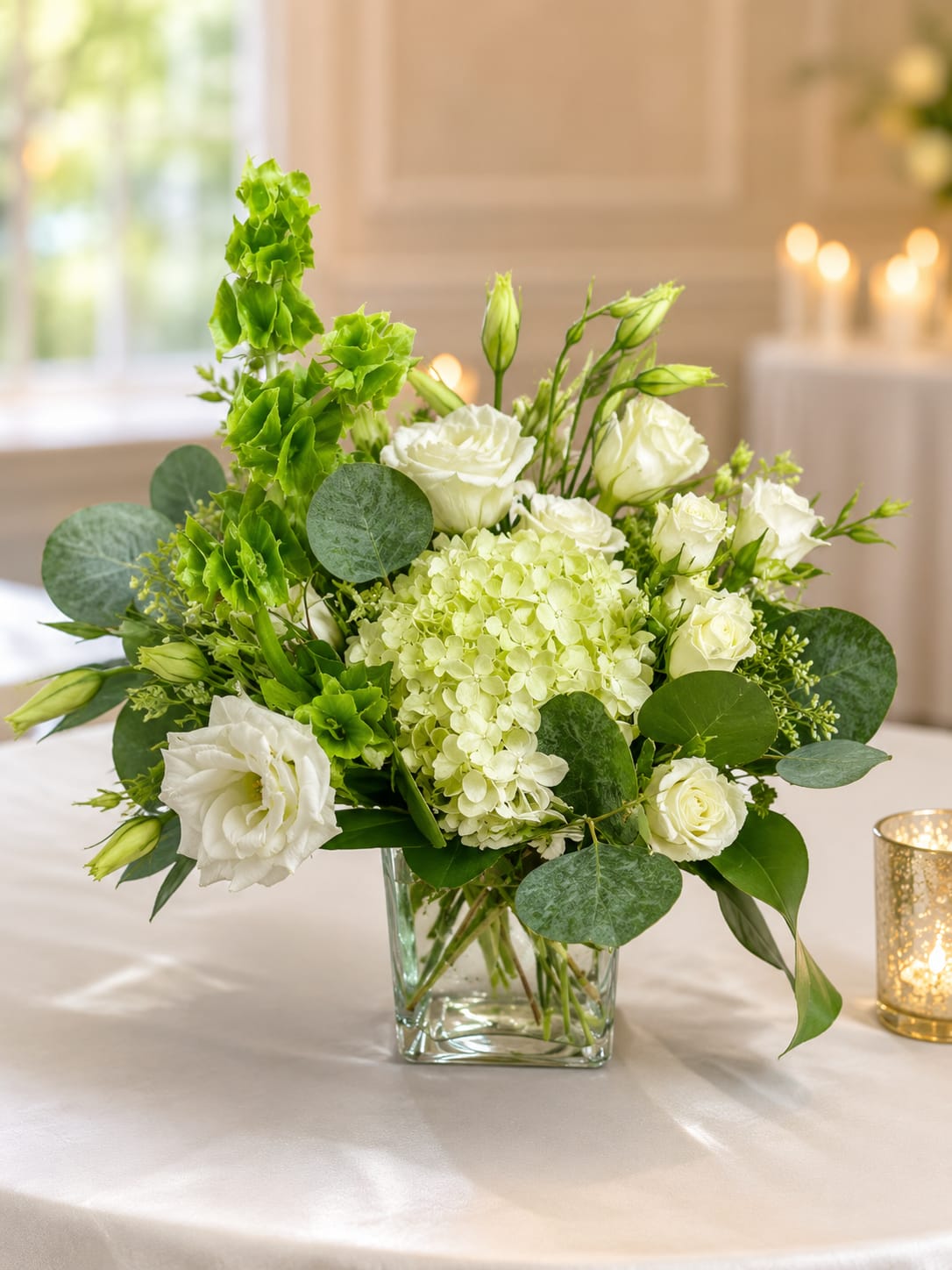 Whimsical White by Petal Perfection Flower Company - Soft green hydrangea forms a plush, inviting base while delicate white lisianthus blooms and buds lend a refined, airy touch-this white and green flower arrangement is an elegant choice for a sympathy arrangement, Mother's Day gift, or a soothing "thinking of you" bouquet. A vertical spray of bright Bells of Ireland creates a sculptural, contemporary line on one side that catches the eye and gives the design movement without overpowering the calm color story.  Arranged in a clear glass cube vase that reveals the clean stems, this compact design fits beautifully on a bedside table, entry console, or office desk. Expect a lush, tactile texture from the hydrangea paired with the soft, ruffled lisianthus and the tall, architectural Bells of Ireland; colors stay in a serene palette of white and green meant to comfort and uplift. Size and stem visibility make care simple-fresh water and a cool spot away from direct sun will help blooms last longer-an ideal gift for birthdays, anniversaries, new baby celebrations, graduations, housewarmings, get-well wishes, weddings, or quiet holiday moments like Easter and Thanksgiving.