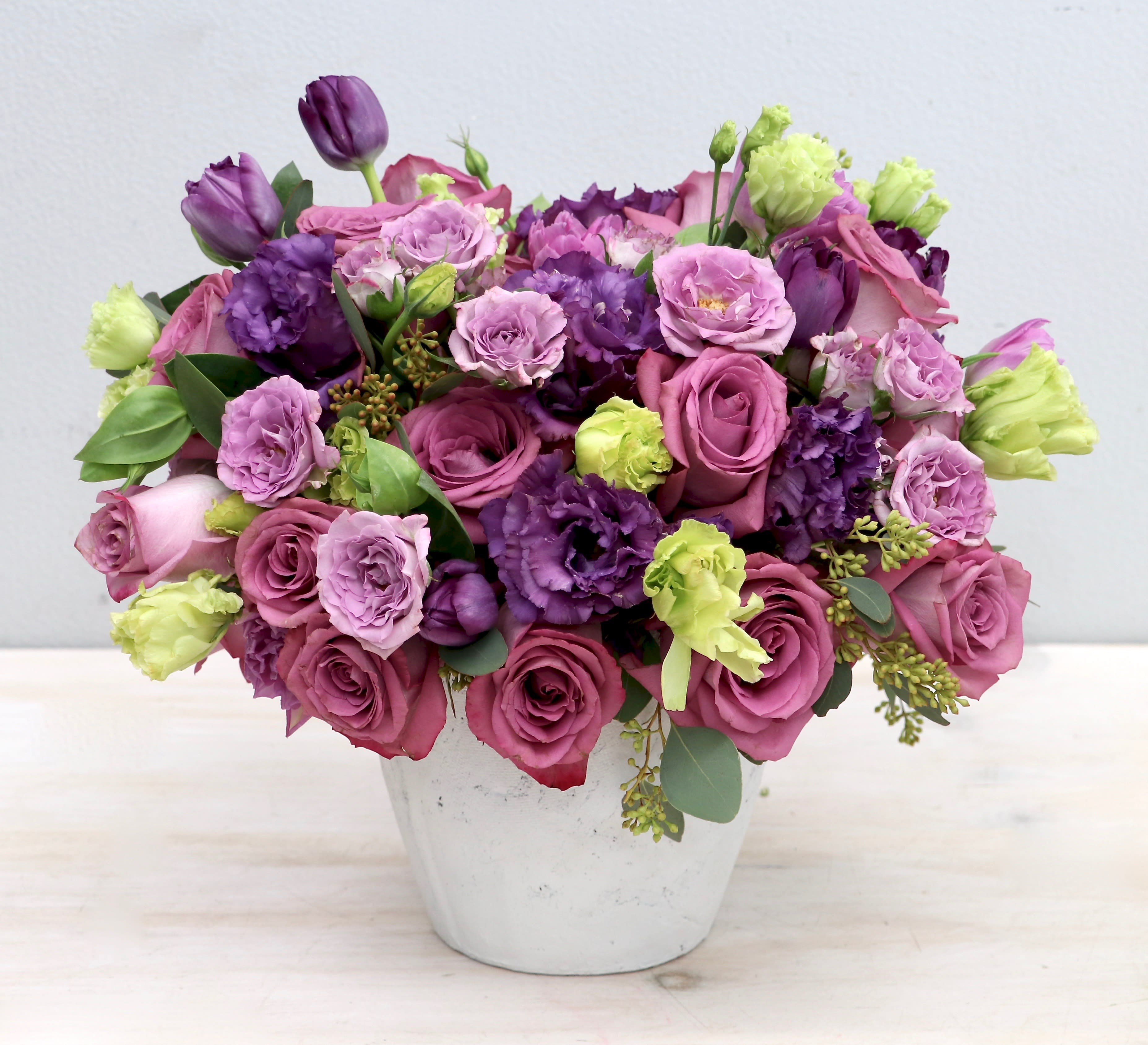 Compact arrangement of purple and pink roses with lisianthus and tulips in a white pot