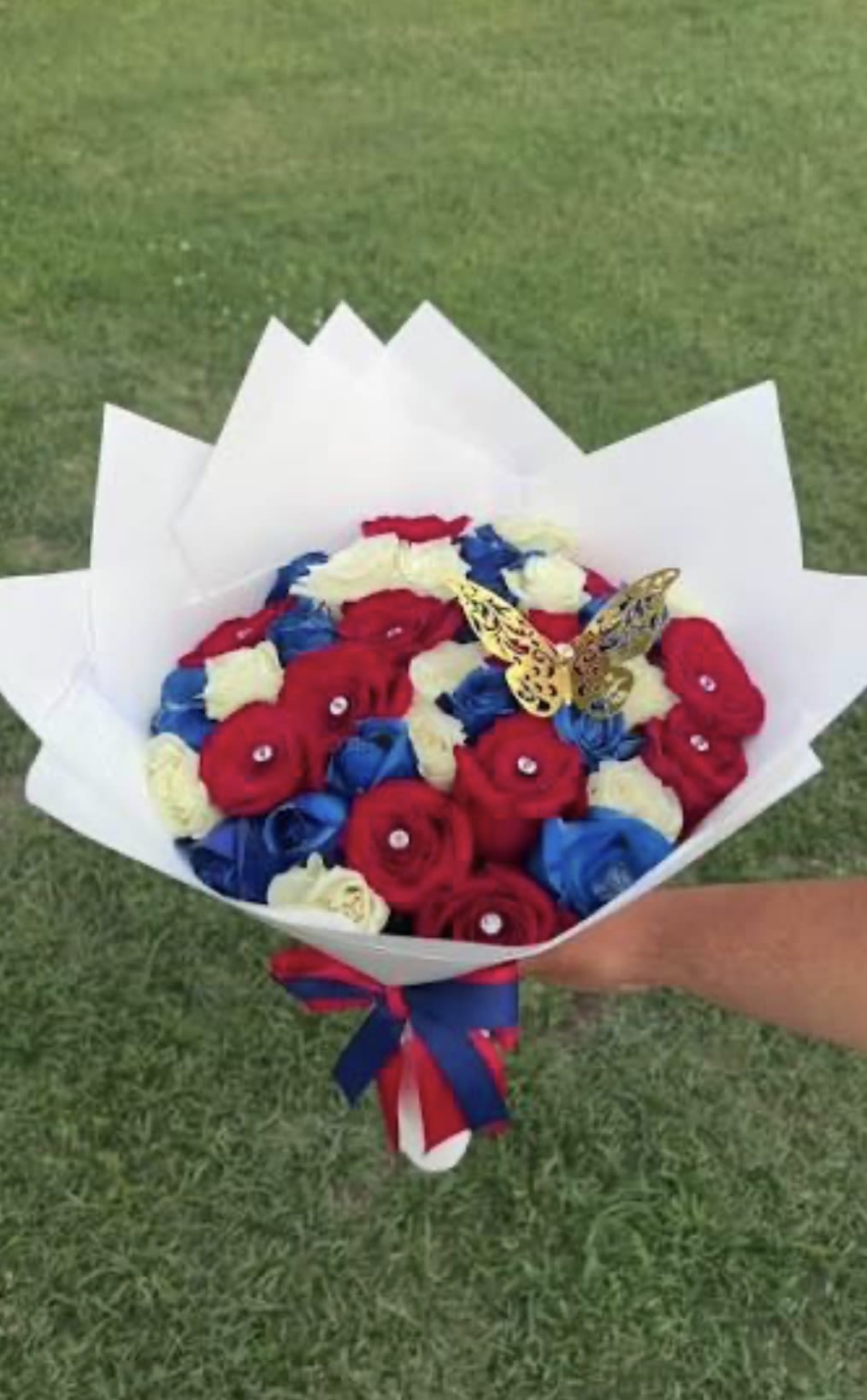 4TH JULY - A boldly patriotic bouquet built from tightly packed red, blue, and white roses, perfect as a 4th of July bouquet or any celebratory arrangement for birthdays, anniversaries, or get-togethers. The rounded, hand-tied shape presents a strong tricolor impact that reads festive at first glance-she'll feel noticed and celebrated the moment the bouquet arrives.  Each bloom is arranged close together to emphasize color and texture; vibrant red, dyed blue, and crisp white roses are wrapped in crisp white paper that frames the saturated petals. A red-and-blue ribbon secures the base and two decorative butterfly picks add a playful, whimsical accent that lifts the design. The arrangement is eye-catching and made to travel well as a gift-ready presentation.  Ideal for patriotic holidays, backyard barbecues, or any moment you want to make someone feel truly celebrated, this bouquet sends a clear, joyful message. Hand-tied and wrapped for immediate gifting-no extra fuss required-so you can add it to a party spread or present it as a bright, memorable surprise.