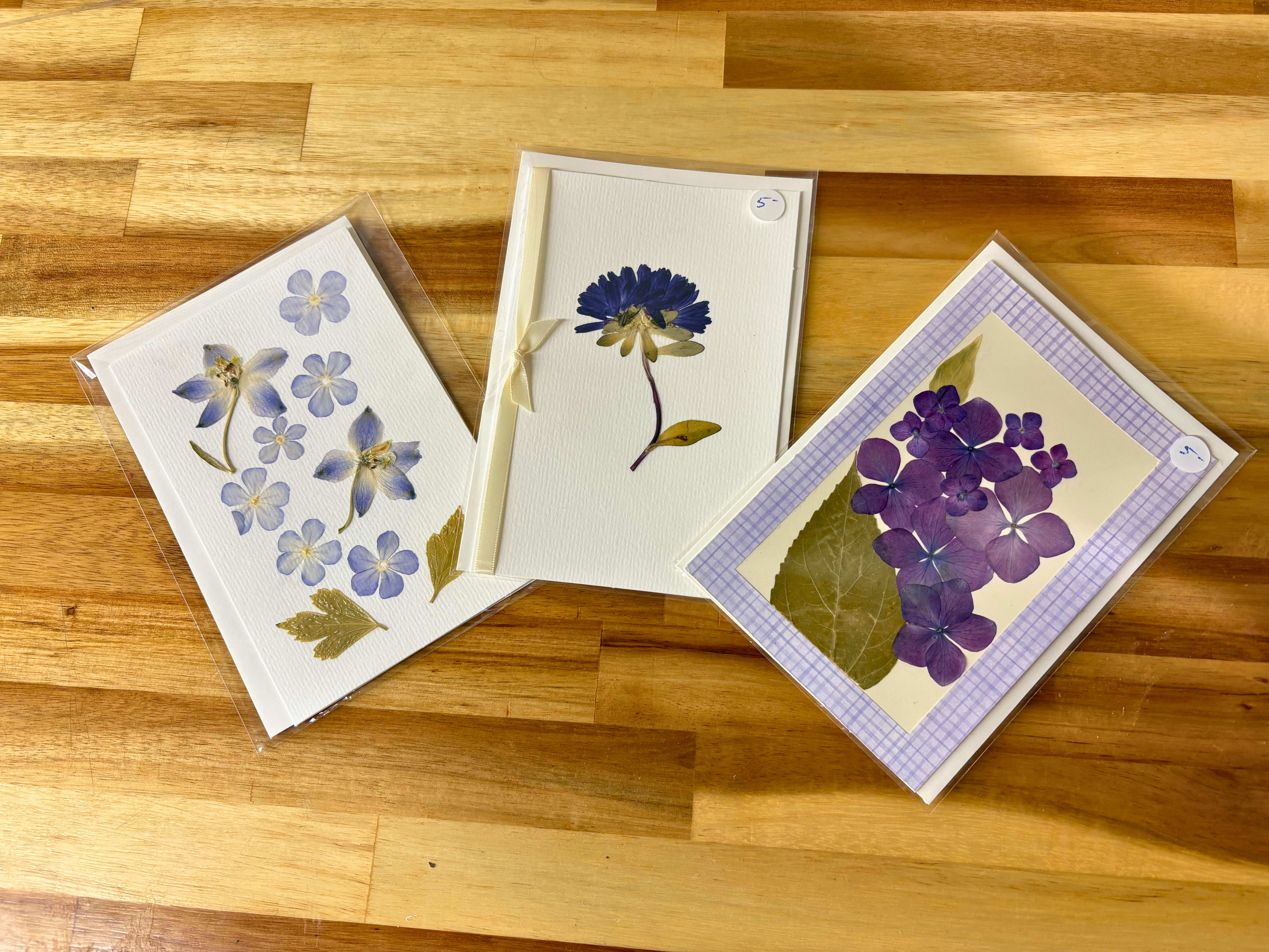 Three pressed-flower greeting cards on a wooden surface.