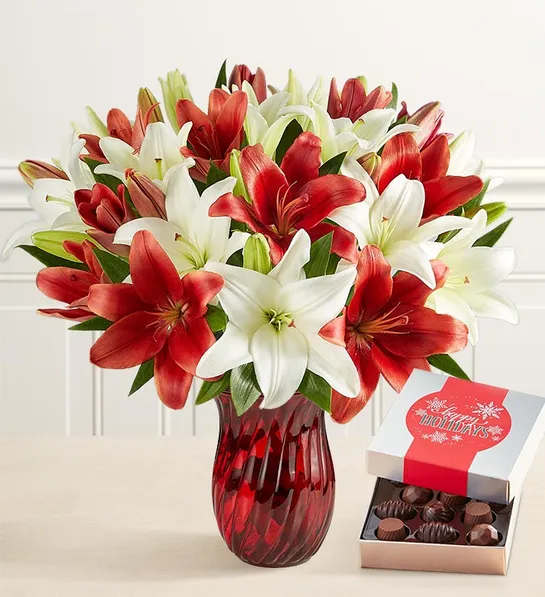 Red and white lilies in a red glass vase beside an open box of chocolates