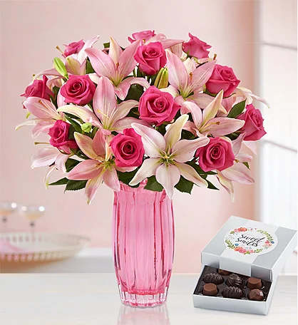 Pink roses and lilies in a pink glass vase beside a box of chocolates