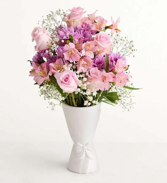 Mom's Garden BQT - Soft pink roses paired with purple daisy-like blooms and delicate pink alstroemeria create a romantic birthday bouquet or anniversary arrangement that feels both feminine and fresh. The pastel mix of pinks, purples and touches of white makes this an ideal Mother's Day gift or spring celebration surprise - the person you're thinking of will feel cherished the moment they see the soft, rounded composition.  Arranged in a tall tapered white vase with a subtle bow detail, the full, rounded bouquet includes airy white filler flowers for lightness and texture. The presentation can be styled on a pale wood tray with a lit candle and round coasters for an elegant home accent; the included white vase and optional wood tray and glass candle holder make it gift-ready and easy to display on a dining table, mantle, or bedside.    Perfect for anniversaries, birthdays, or simply to say "I'm thinking of you," this arrangement arrives ready to brighten a room and lift spirits. The vase sizing is ideal for tabletops and entryway consoles; to keep blooms looking their best, place away from direct heat and refresh the water every few days.