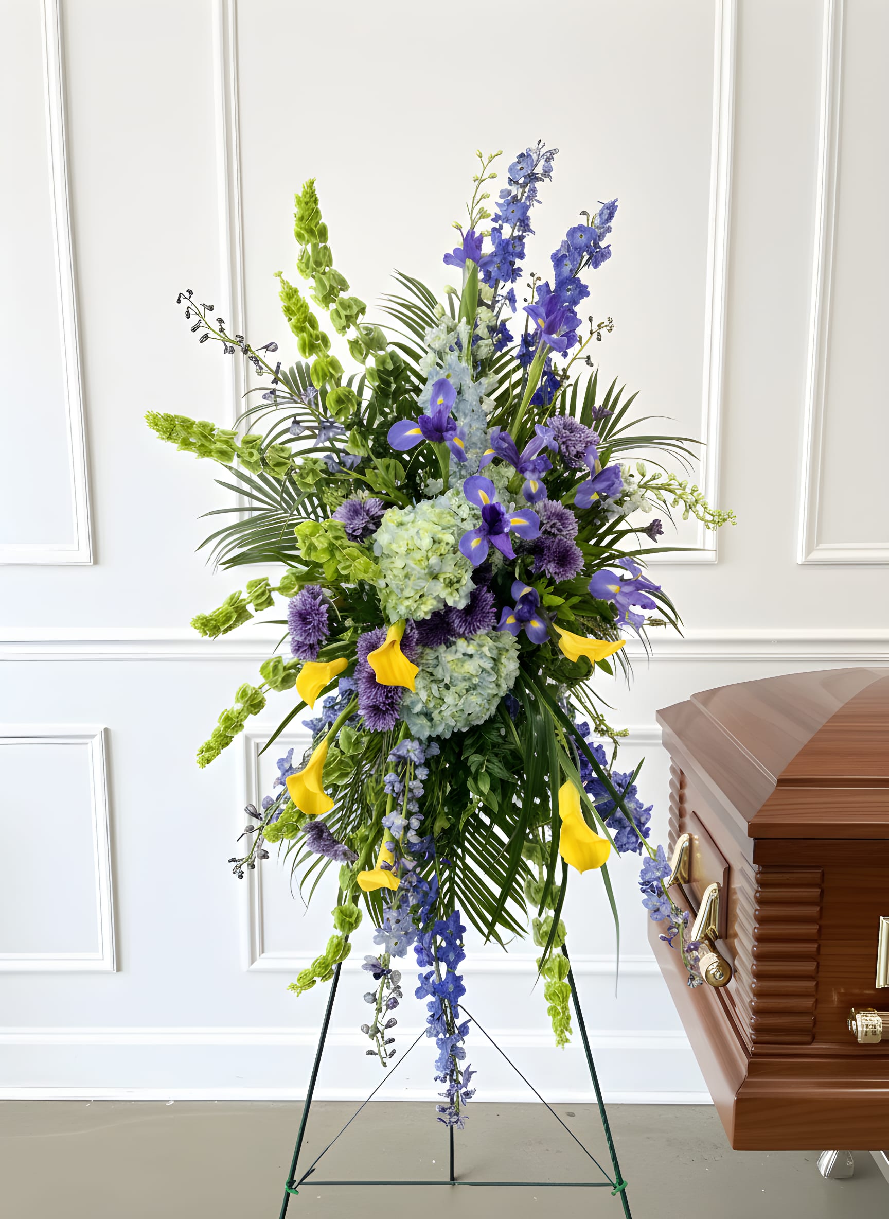 Tall funeral spray with blue, purple, green, and yellow flowers beside a casket