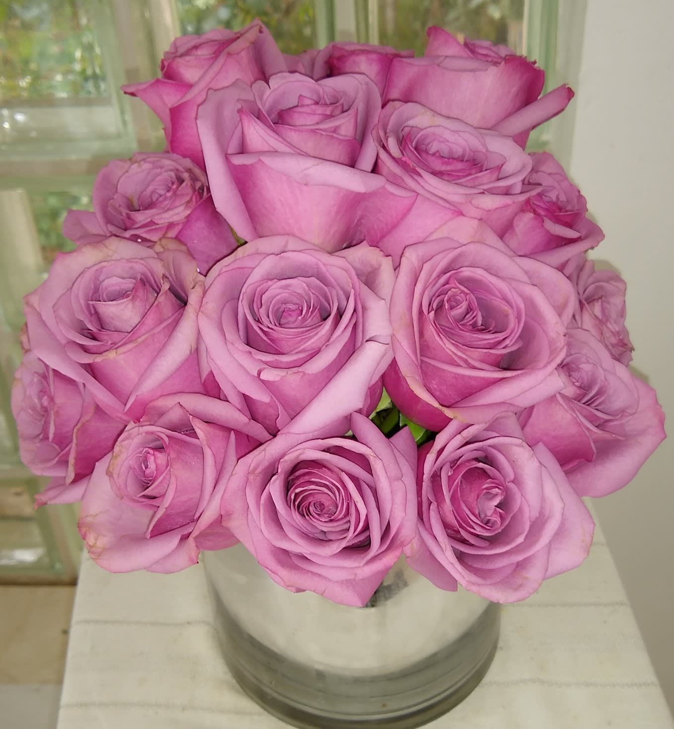 Enchanted Love - Lavender roses cut tight and compact in a low cylinder.   Perfect for anyone's desk, end table or as a centerpiece. Approximate dimensionts:  10"D X 10"H