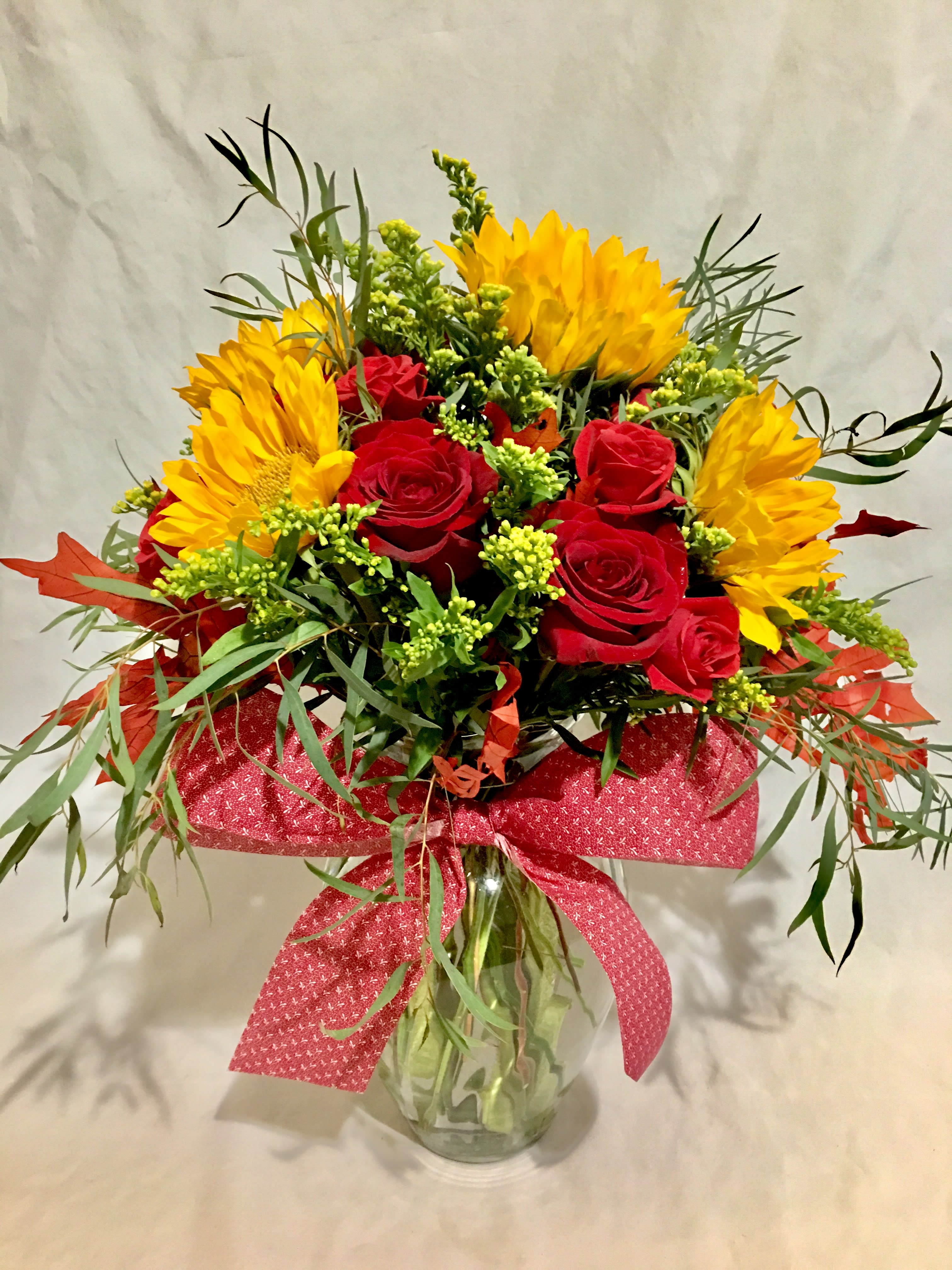 Clear vase arrangement with yellow sunflowers and red roses accented with a large red bow.