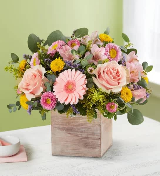 Low arrangement of pink roses, gerbera daisies, and yellow button blooms in a rustic wooden cube vase.