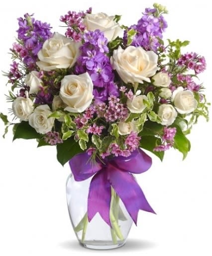 ENCHANTED COTTAGE ARRANGEMENT - As enchanting as coming across a hidden cottage in the countryside! Lush white roses and purple stock mix with delicate lavender waxflower and green pitta negra for an elegant English garden look. The decorative bouquet is finished with a pretty satin ribbon.
