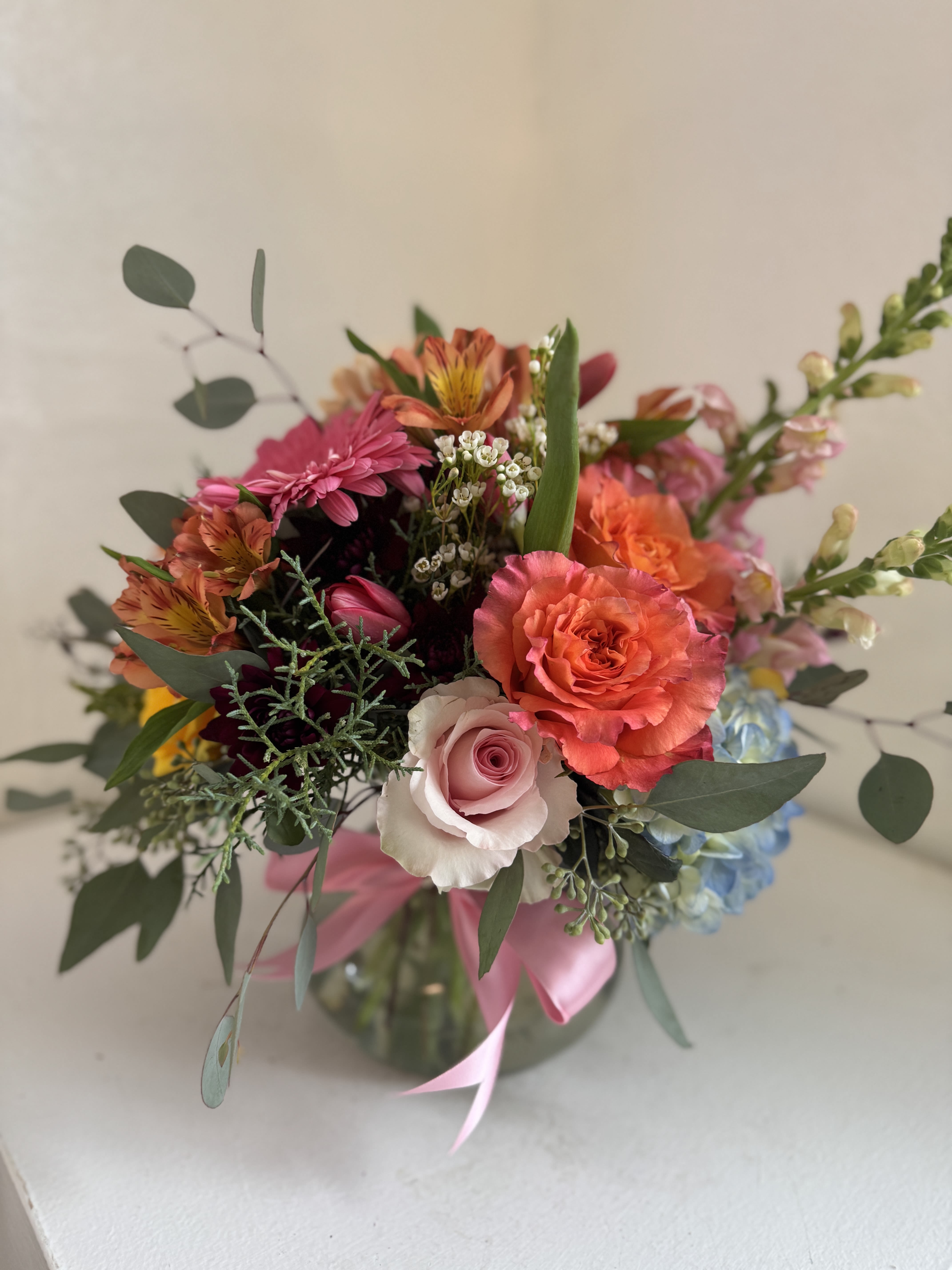 Mixed bouquet of pink, orange, and blue flowers in a glass vase with a pink ribbon