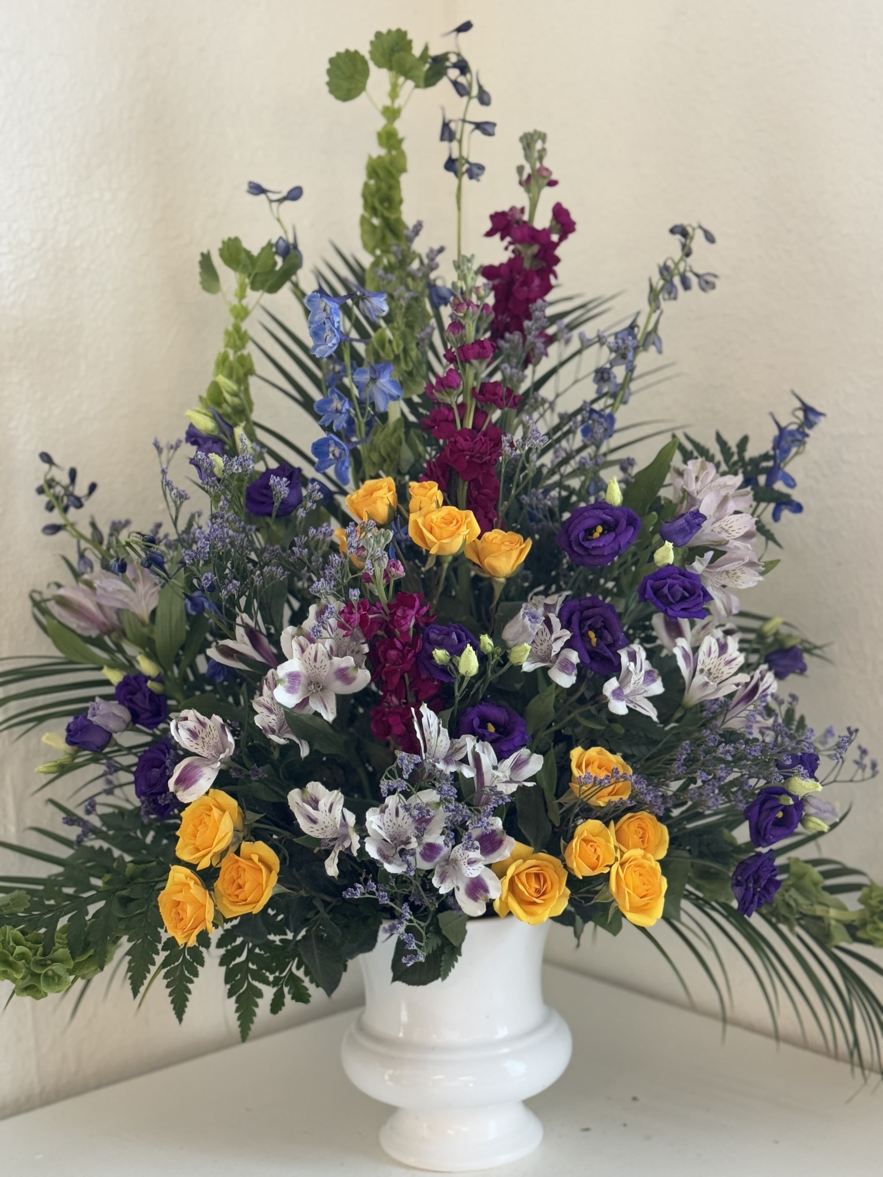 Tall mixed floral arrangement in a white pedestal vase with yellow, purple, and white blooms