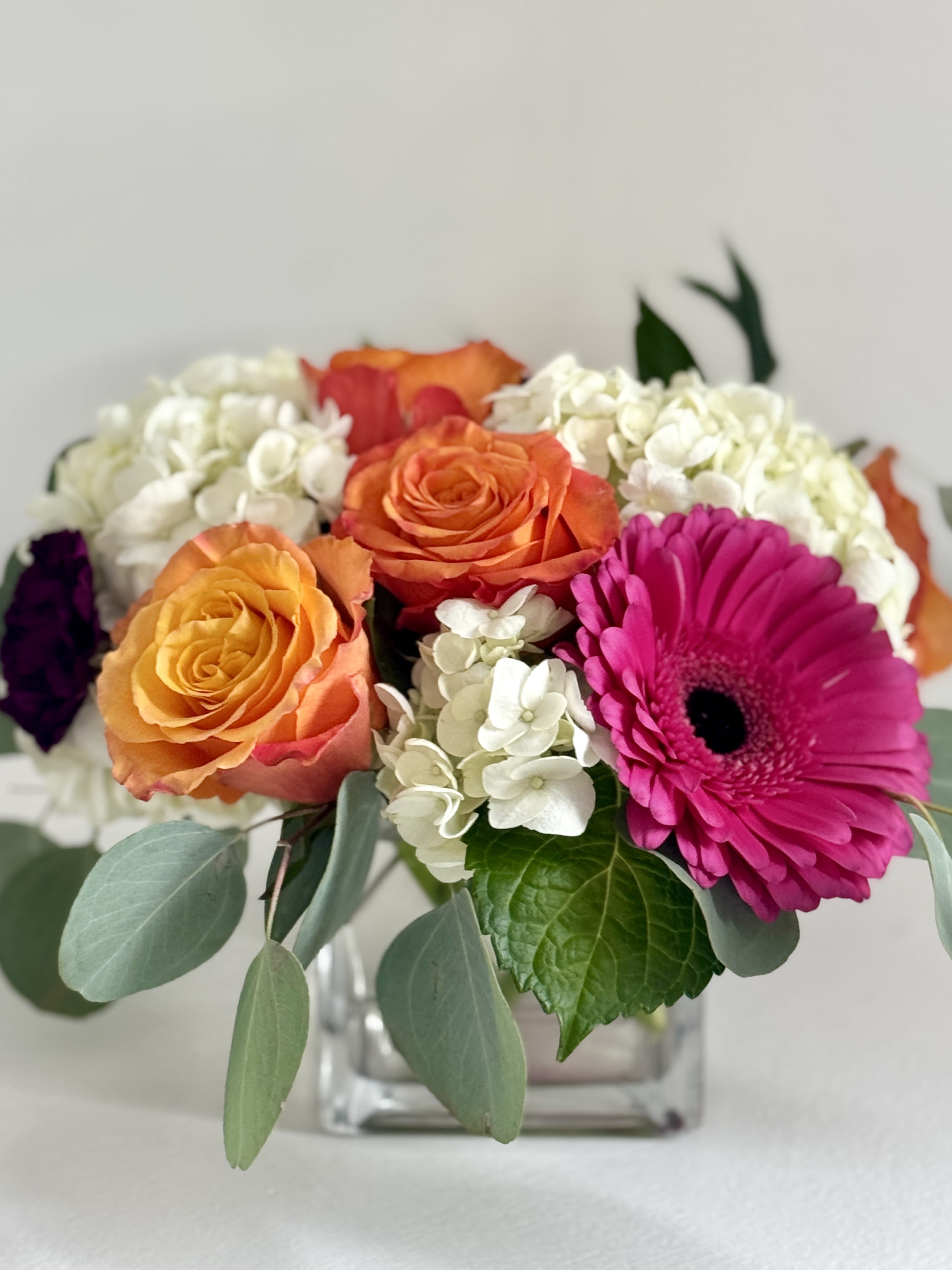 Bouquet of orange roses, white hydrangeas, and a pink gerbera daisy in a glass vase