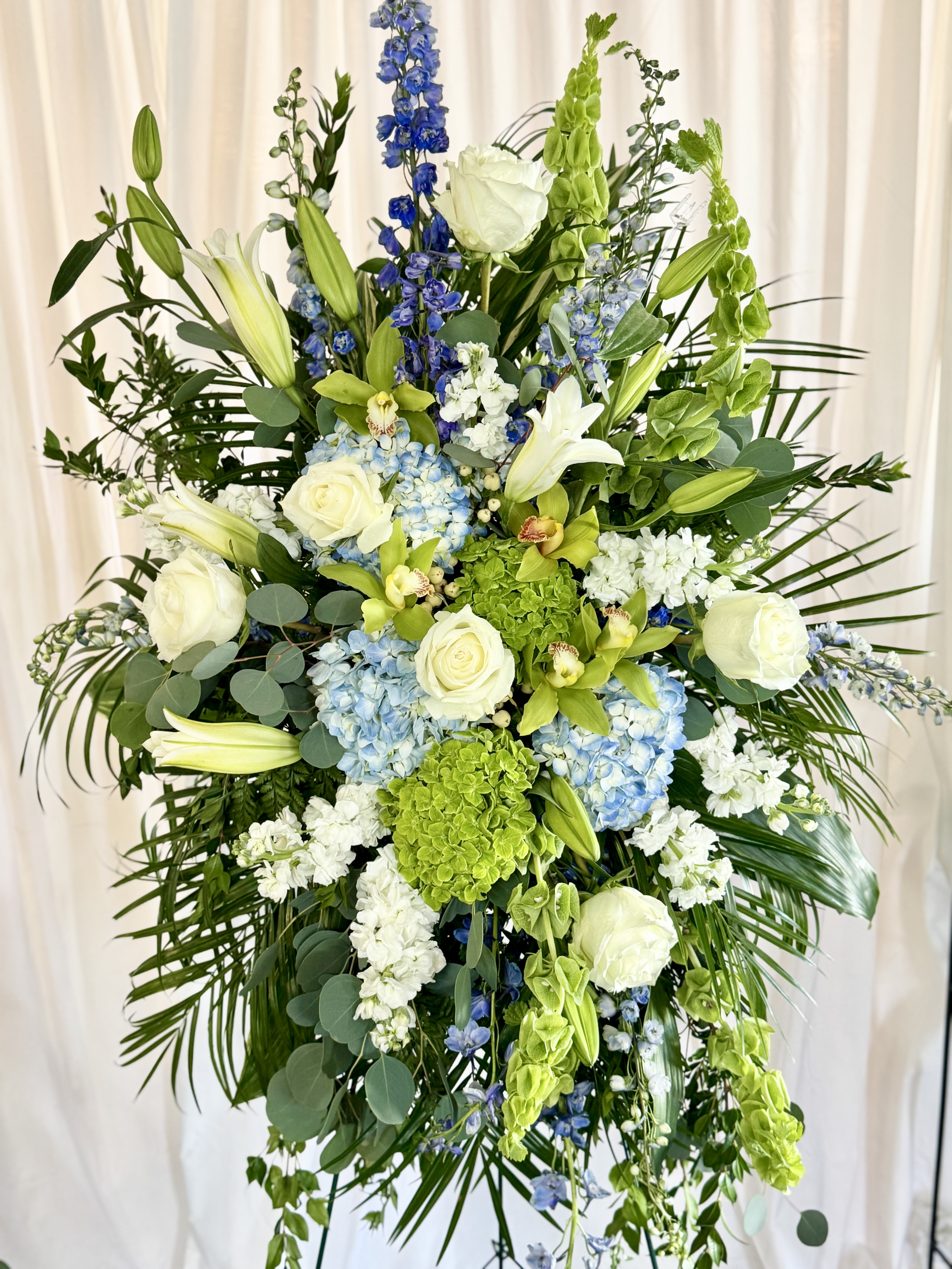 Tall floral spray with white roses, lilies, blue hydrangeas, and green blooms