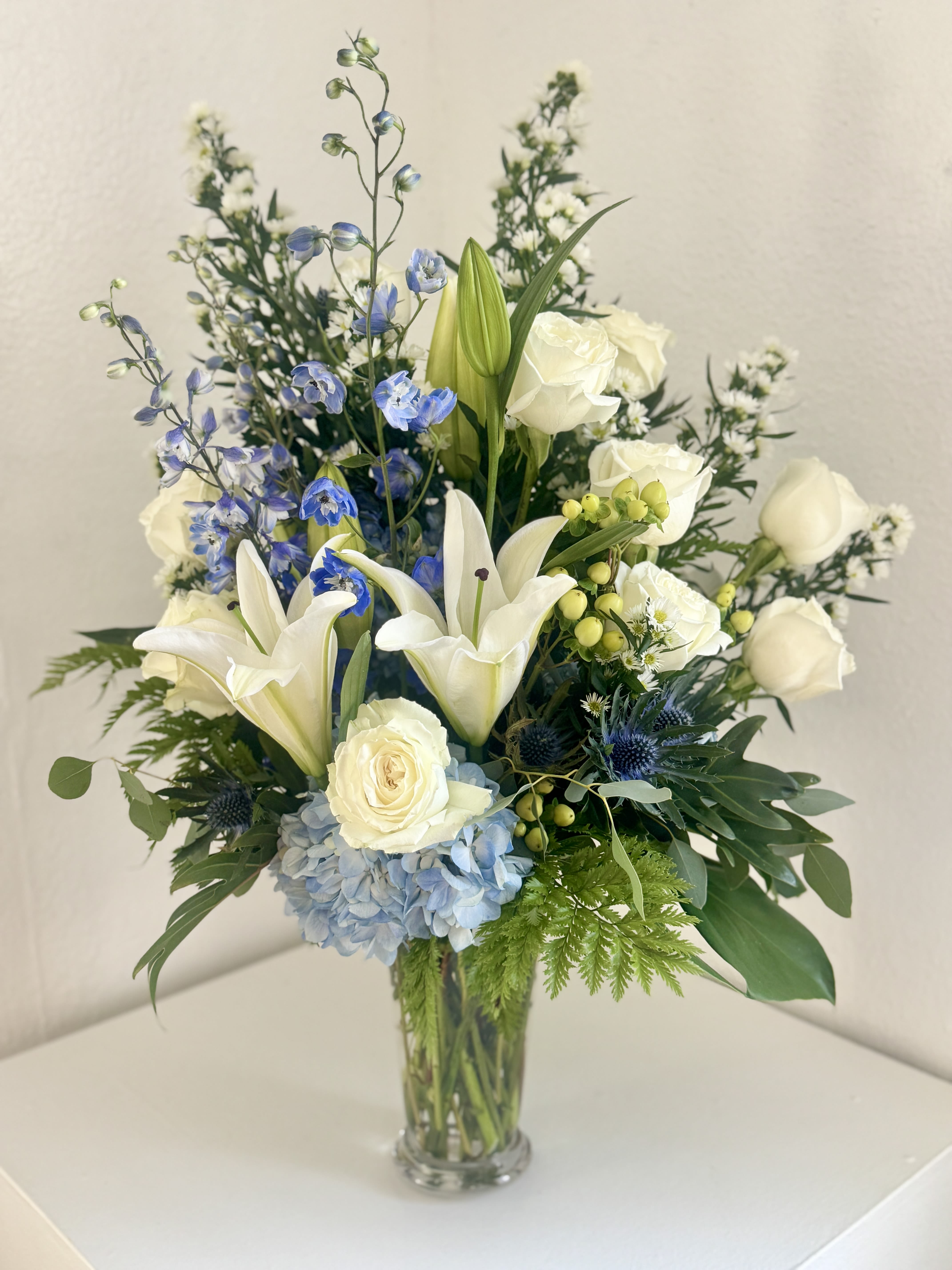 White lilies, roses, and blue flowers in a clear glass vase