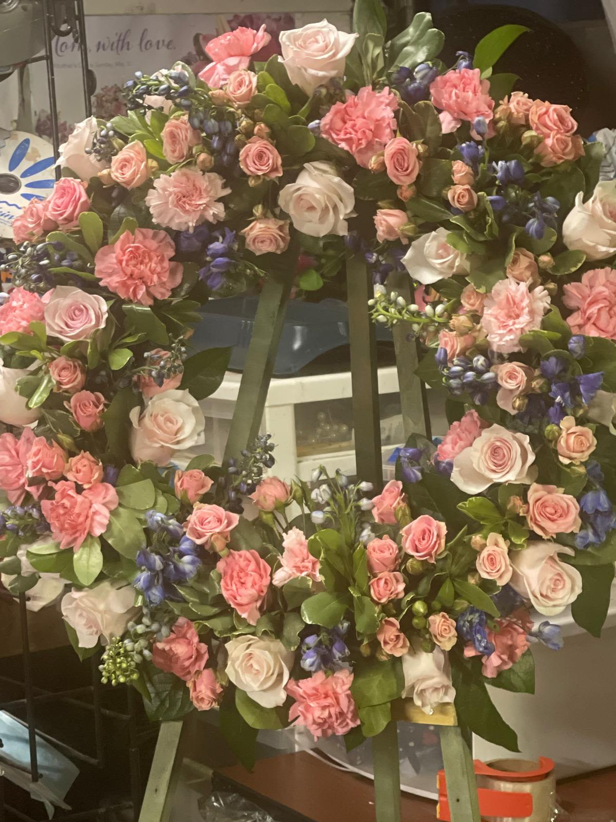 Standing wreath of pink roses and carnations with blue accent flowers on a green easel.