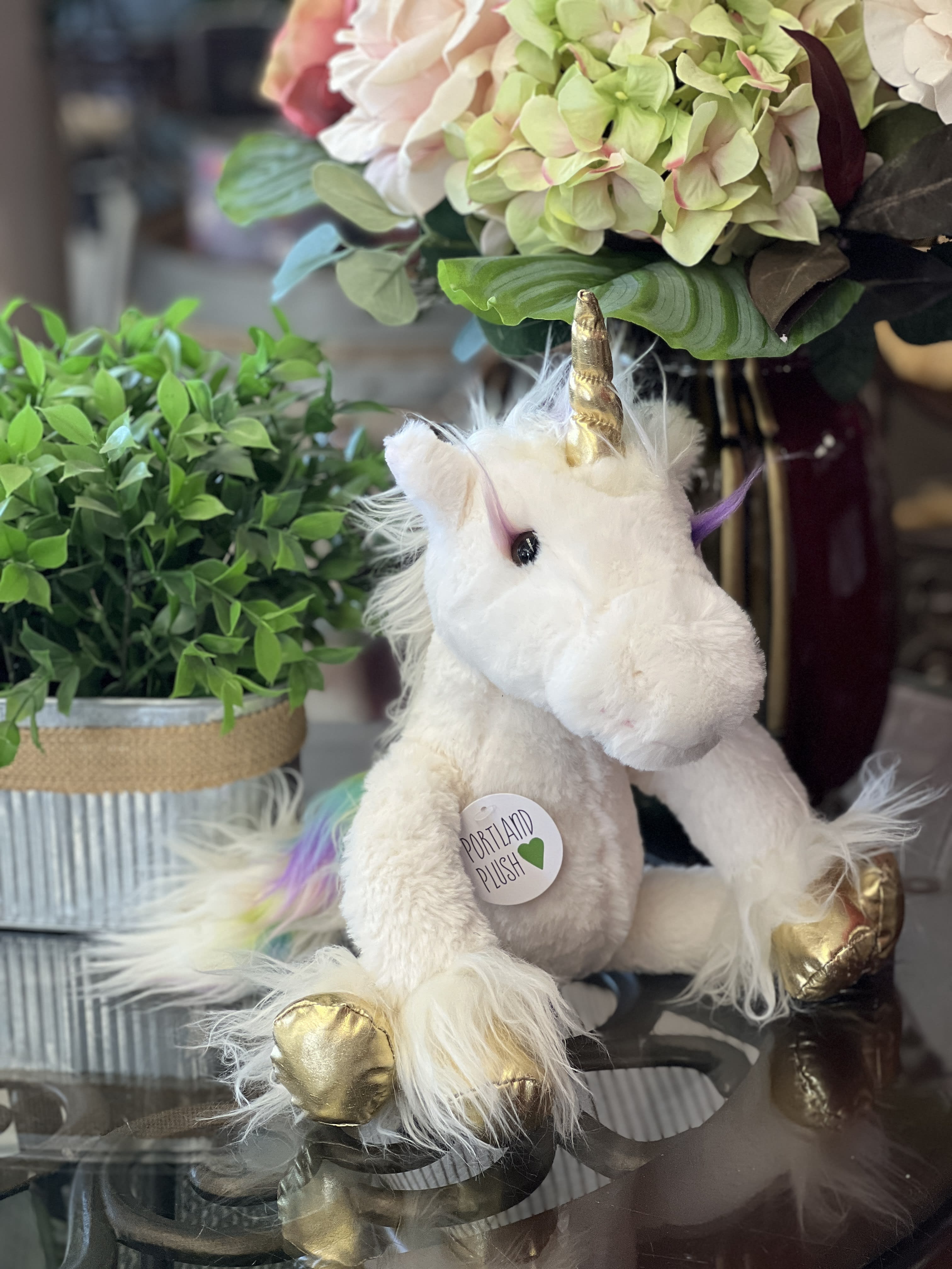Plush unicorn with gold horn and hooves beside potted greenery and hydrangeas