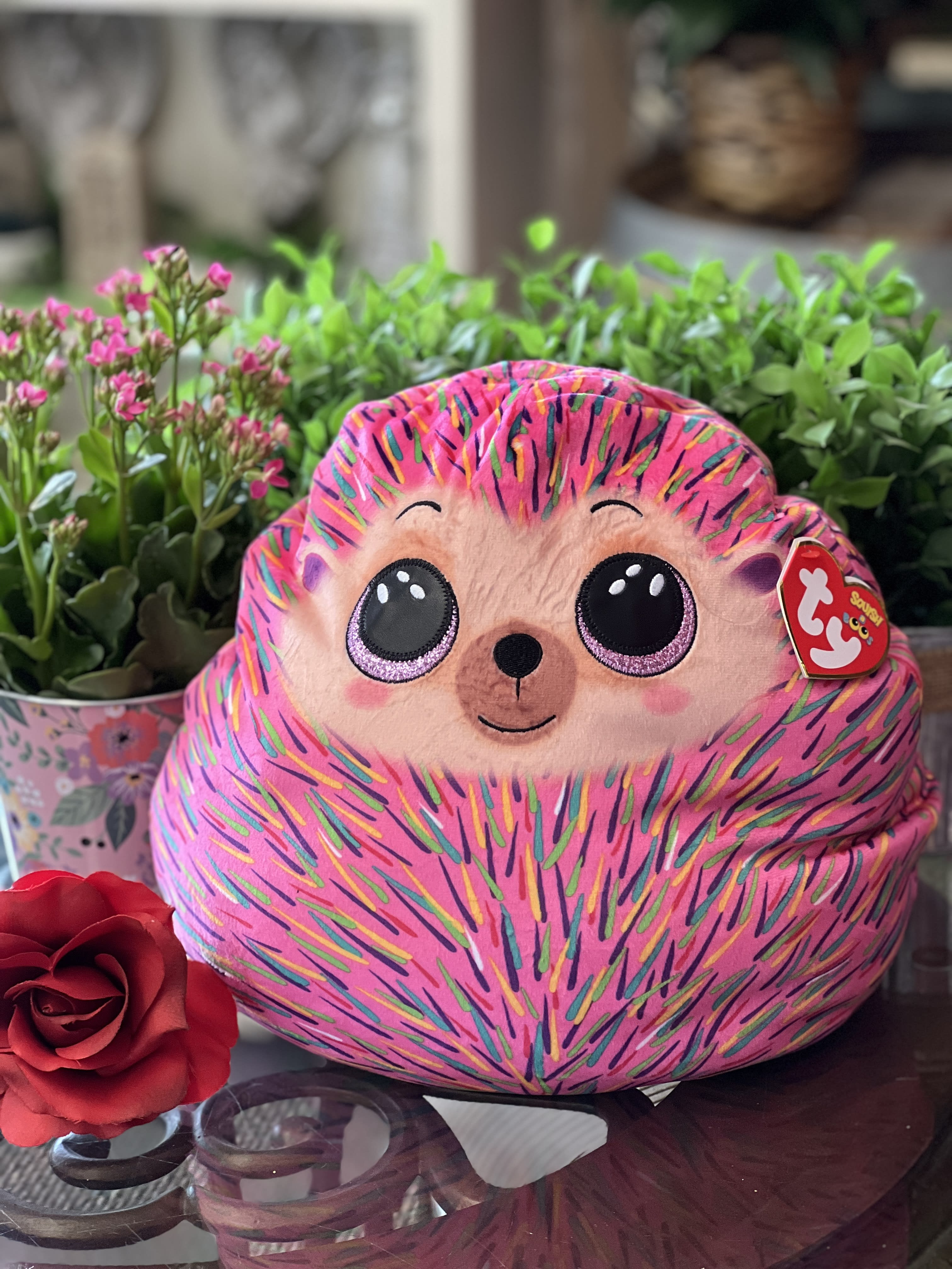 Pink plush hedgehog toy with a red rose and potted flowers behind it