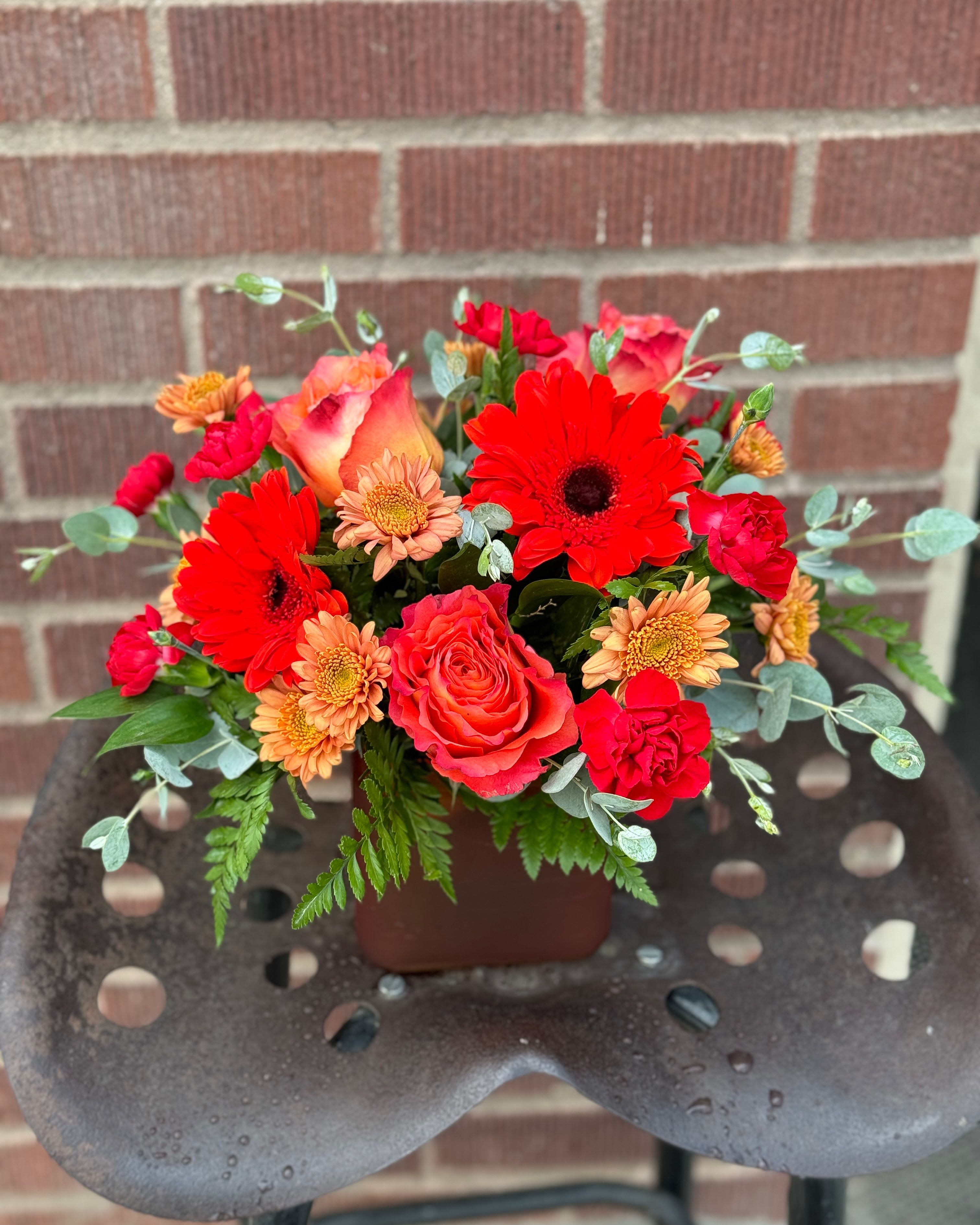 Mixed bouquet of red, coral, and peach flowers in a brown container