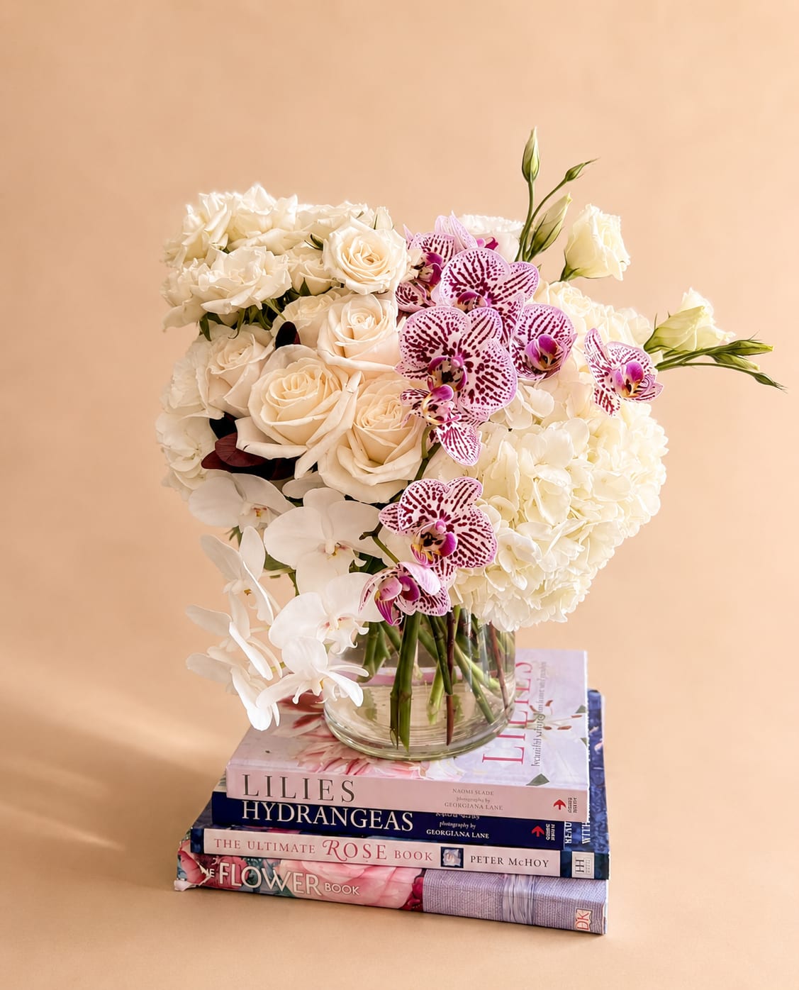 White roses, hydrangeas, and orchids arranged in a glass vase on stacked books