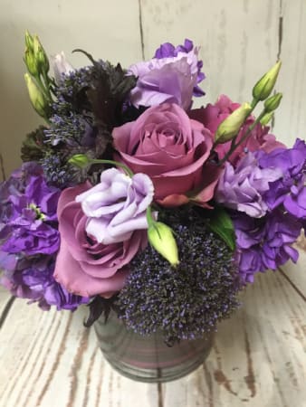 Compact arrangement of purple and lavender roses and blooms in a clear glass cylinder vase