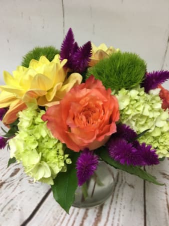 Low arrangement of orange rose, green hydrangea, yellow blooms and purple flowers in a clear glass vase