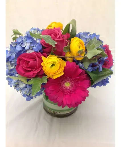 Bright bouquet of pink roses and gerbera daisy, yellow ranunculus, and blue hydrangeas in a glass vase