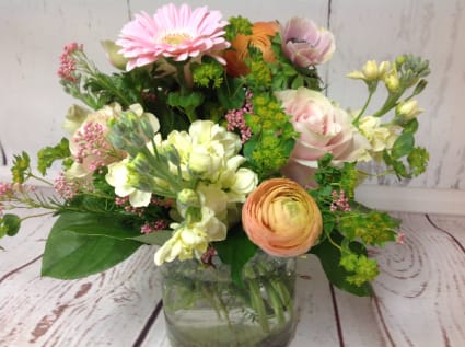 Mixed arrangement of pink, peach, and cream flowers in a clear glass vase on a wood surface