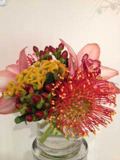 Compact arrangement of pink lilies, red pincushion protea, yellow blooms, and berries in a clear glass vase.