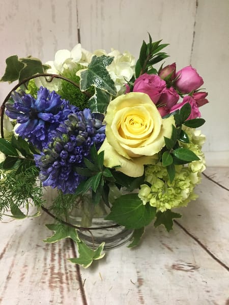Short glass vase arrangement with yellow rose, pink roses, blue hyacinth, and white blooms
